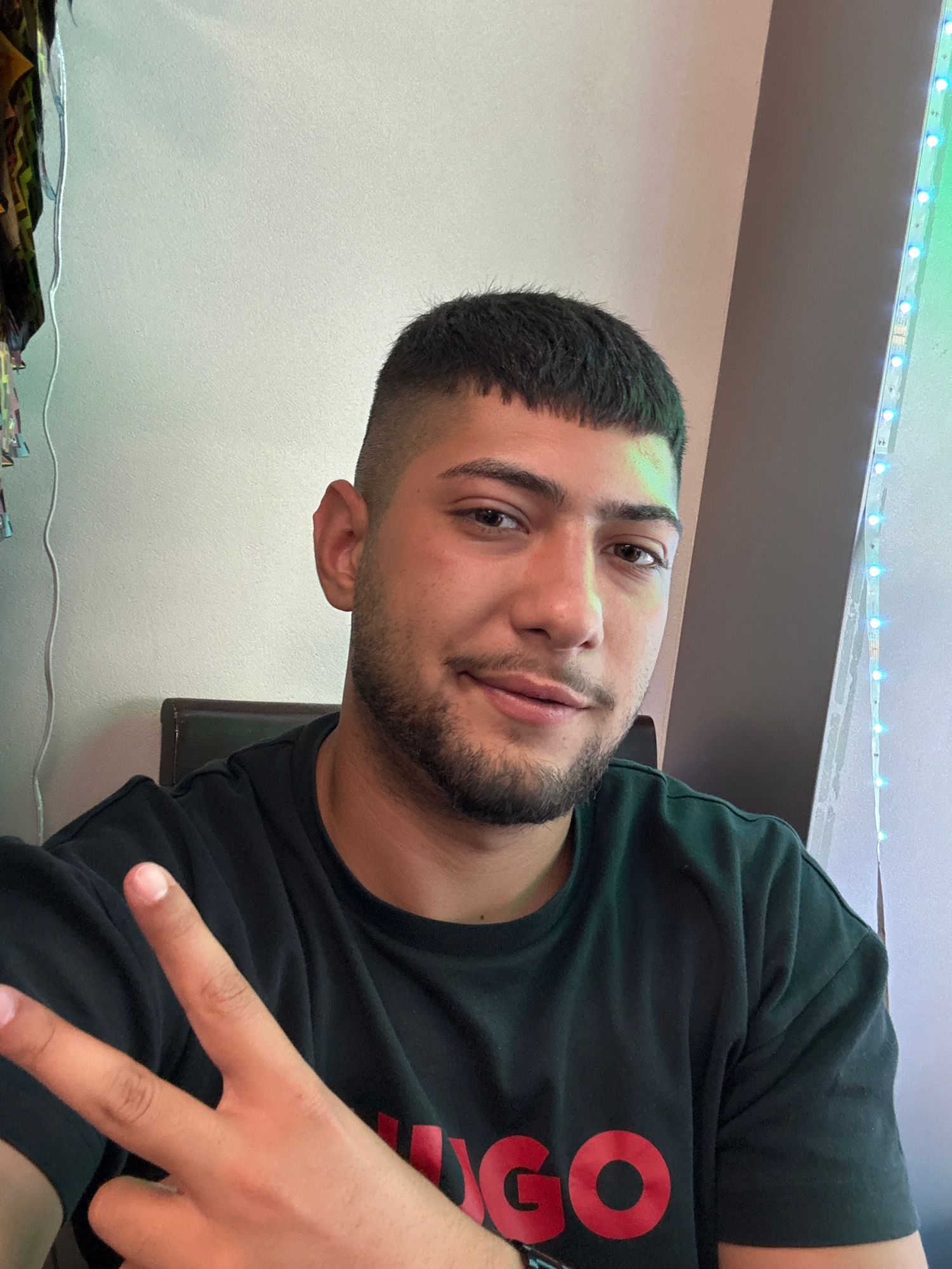 A young man with a short haircut and a beard is looking at the camera, holding up two fingers in a peace sign. He is wearing a black t-shirt with red lettering. The background is a wall, with a string of blue LED lights on the right side. The image does not provide any information about the location in Liestal, Switzerland.