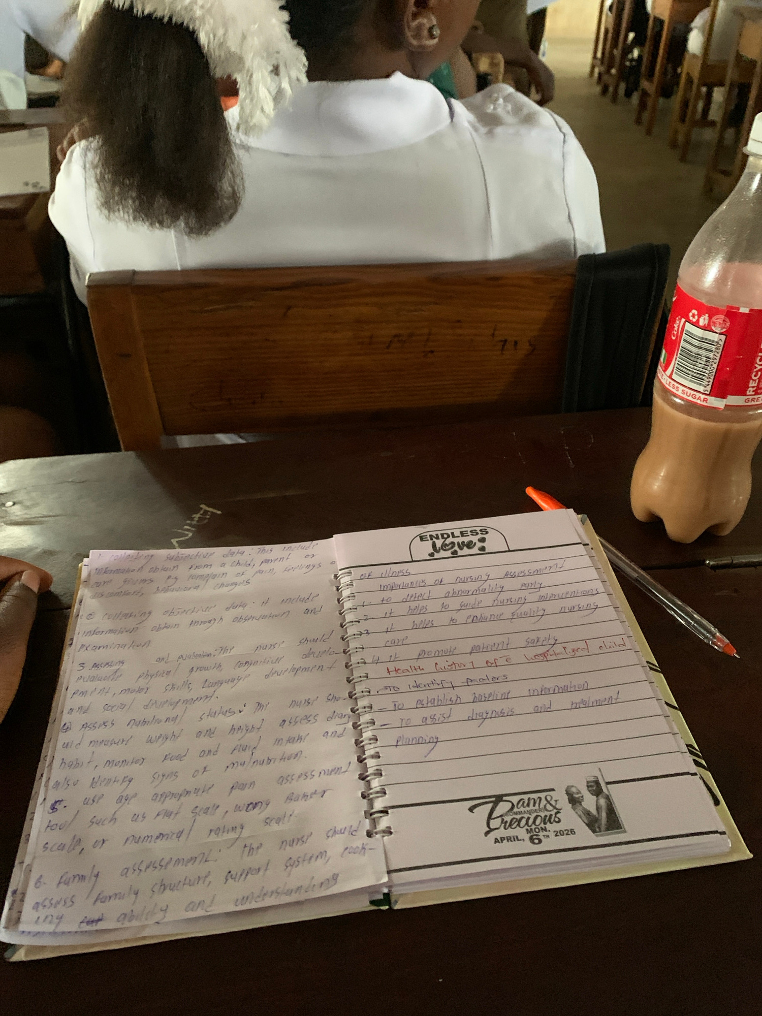 The image shows a close-up of a notebook with handwritten notes and a pen lying on a wooden surface, likely a desk. The notebook has a decorative cover with "Dam & Precious" and a date of "April, Nov. 2026". The handwritten text appears to be related to nursing or healthcare, with headings like "Importance of Nursing Assessment" and "Family Assessment".
In the background, a person's head and shoulders are visible, with dark curly hair. They appear to be wearing a white uniform, suggesting a medical or educational setting. A partially visible plastic bottle, with a red label that says "Coke" and "LESS SUGAR", is also in the frame, suggesting a casual or informal environment, possibly during a break. The lighting suggests it could be daytime, indoors. The overall scene depicts a student or professional taking notes, likely in a learning or work environment, with personal items present. The inscription "Jos, Nigeria" provides a specific geographical context for this scene.