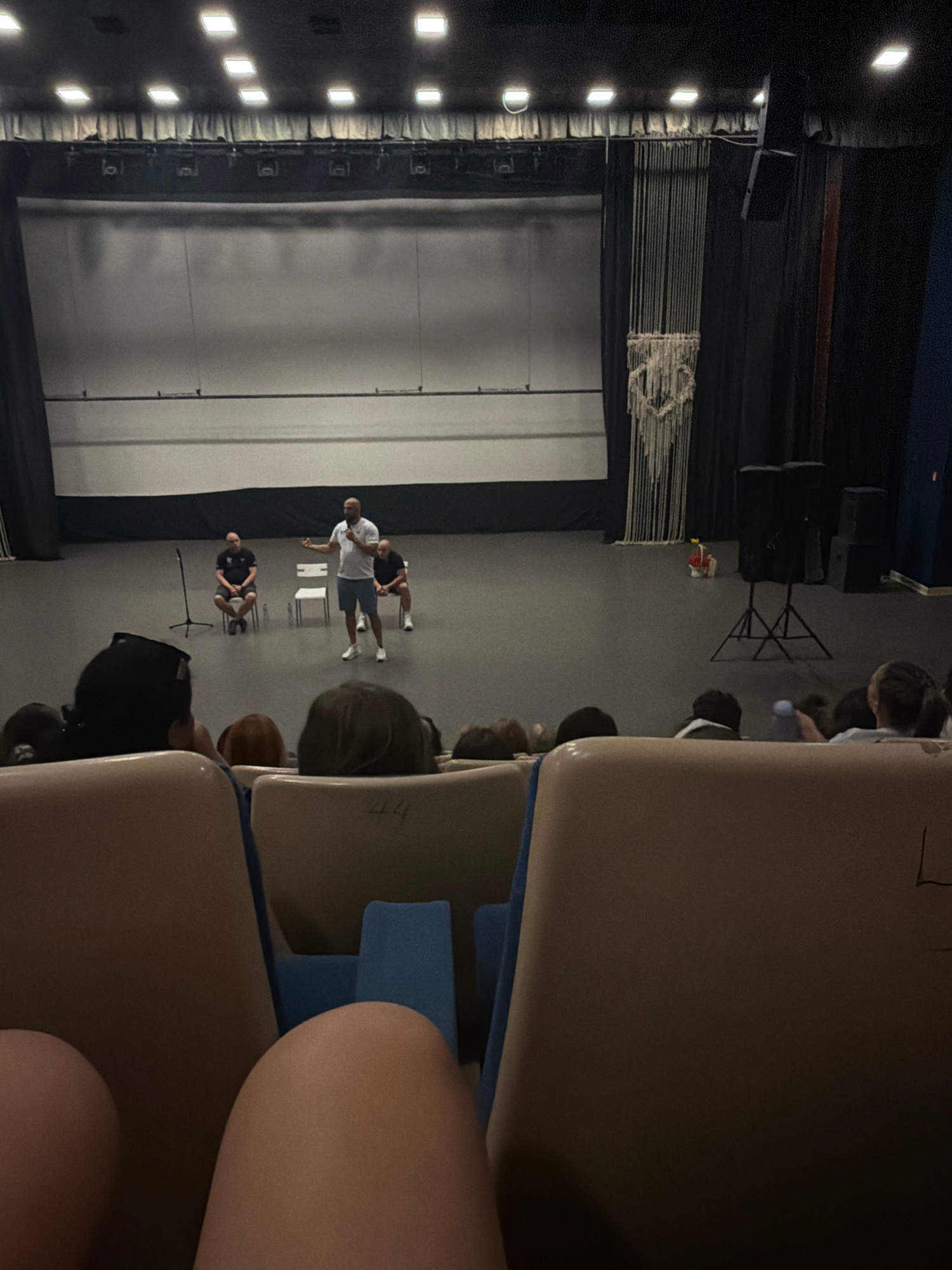 In an indoor performance space, possibly a school auditorium in Blagoevgrad, Bulgaria, a presenter stands on a stage addressing an audience. The stage features a large, plain backdrop and is equipped with microphones and chairs. The presenter, a man in a white polo shirt and blue shorts, gestures as he speaks. Two other men are seated on the stage, one in a black t-shirt and the other in a dark shirt. The audience is seated in rows of chairs, with some members visible in the foreground. The lighting suggests it is an event taking place during the day or with artificial stage lighting.