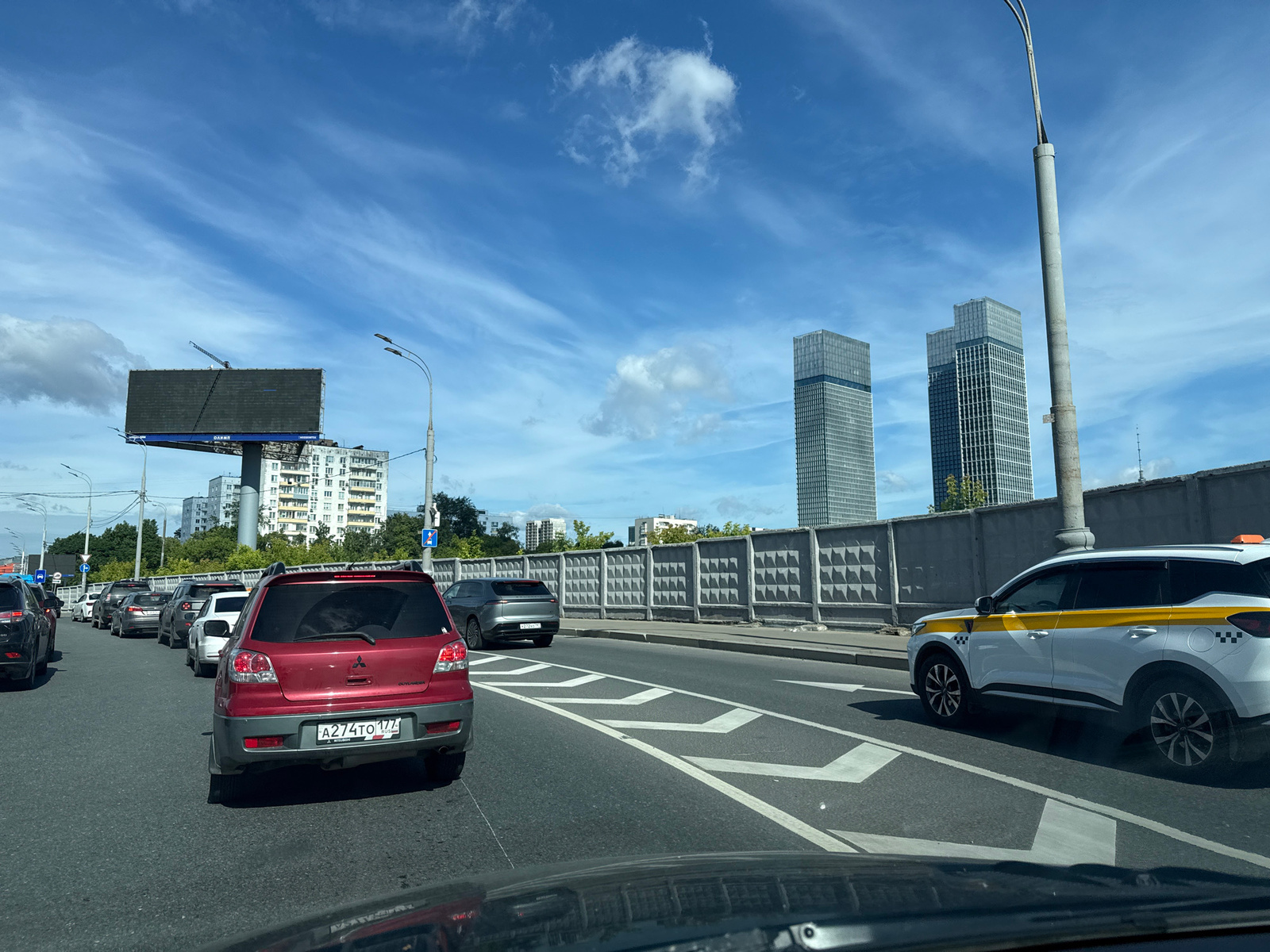 This is a medium shot, taken from inside a car, looking forward on a highway in Moscow, Russia. The sky is blue with wispy white clouds. In the foreground, there are several cars in traffic. The closest car is a red Mitsubishi Outlander with a Russian license plate. To the right, a white taxi with yellow stripes is visible. In the background, there are tall, modern glass buildings, a large billboard, and a concrete fence. Trees and apartment buildings are also visible in the distance. The road has lane markings and is carrying moderate traffic.