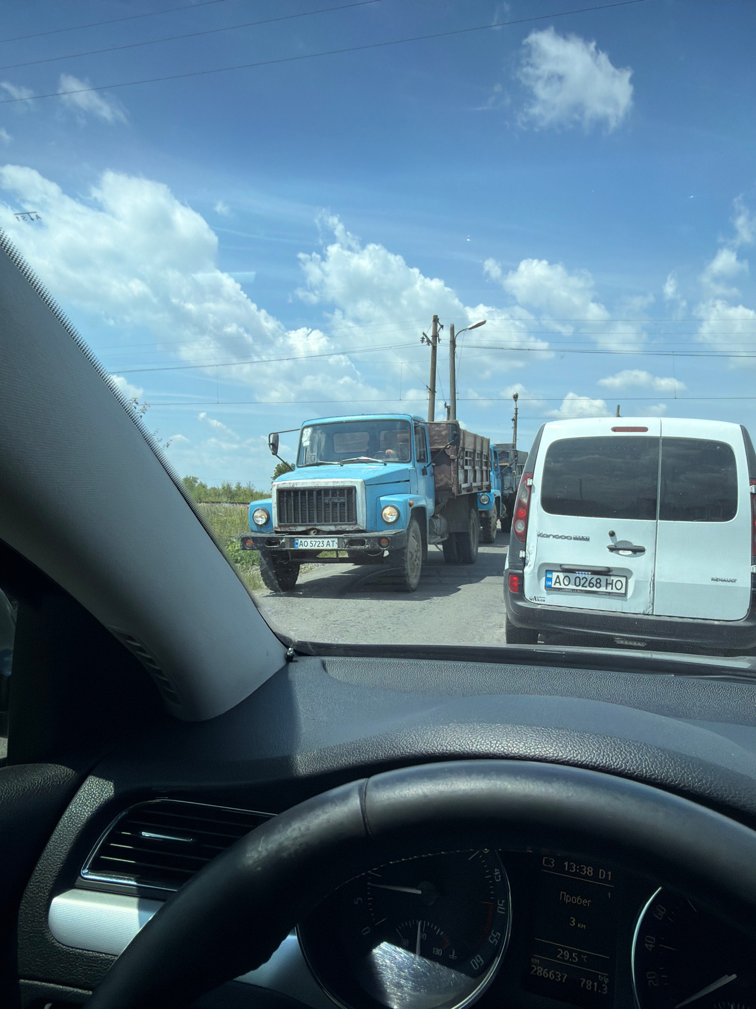 The image shows a view from inside a car, looking forward through the windshield. The car is stopped in traffic on a sunny day. In front of the car, there is a white van, and further ahead, a line of blue trucks. The first blue truck, a flatbed with wooden sides, has the license plate AO 5723 AT. The white van has the license plate AO 0268 HO. The background consists of a blue sky with scattered white clouds, utility poles with wires, and vegetation. The location is identified as Tarnivtsi, Ukraine, based on the provided context. The dashboard of the car is partially visible in the foreground, showing a speedometer and a digital display with time, temperature, and mileage.