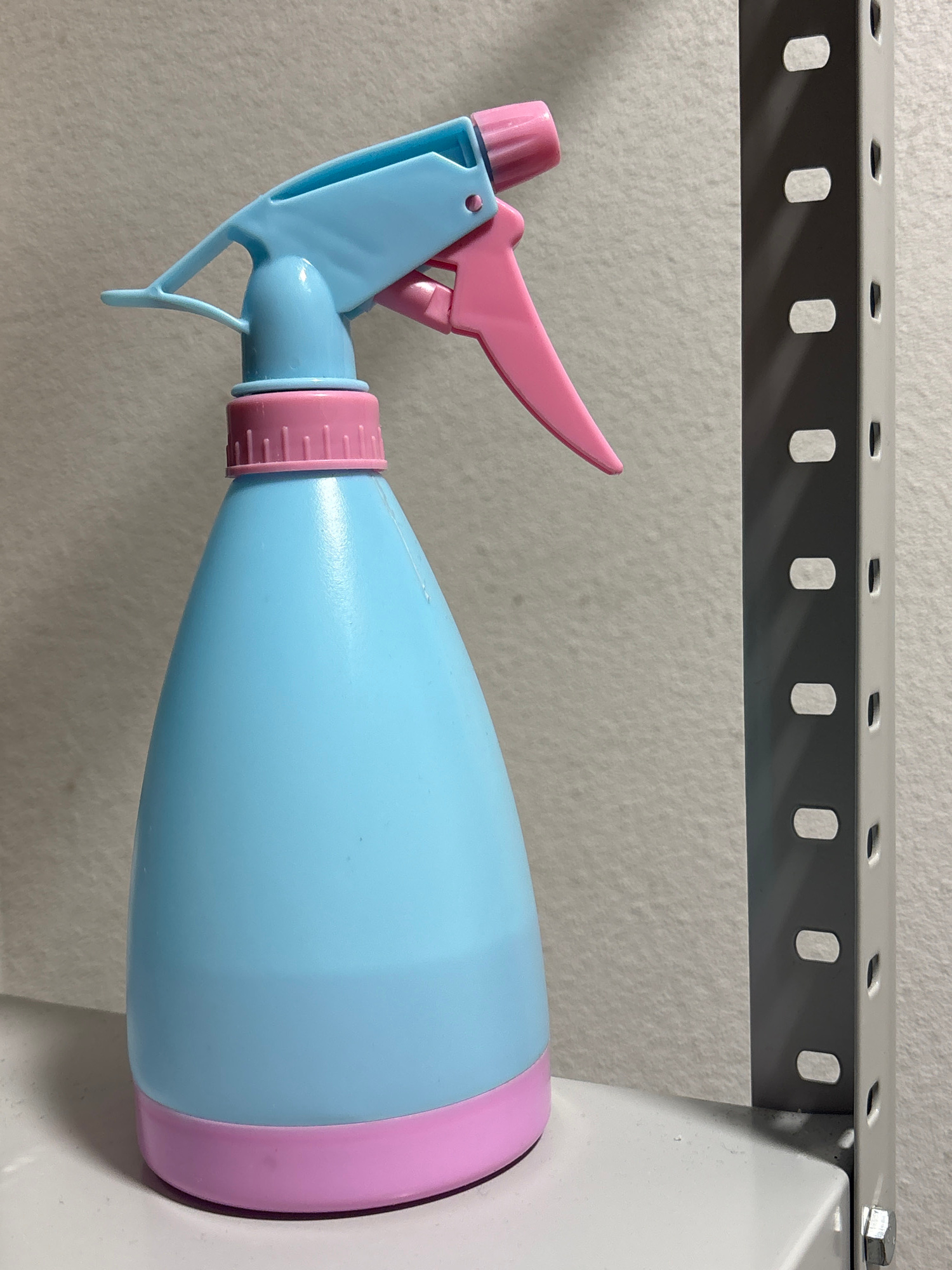 An eye-level, medium shot captures a blue and pink spray bottle positioned on a white shelf. The spray bottle, made of plastic, features a blue bulbous body with a pink base and a pink threaded cap. The spray mechanism is a combination of blue and pink plastic, with a pink trigger. To the right of the spray bottle, a gray metal shelf support with evenly spaced slots is visible, casting a shadow on a textured, light-colored wall behind the bottle. The shelf the bottle rests on is also white. The scene is lit with bright, direct lighting, highlighting the texture of the wall and the reflective surfaces of the objects. The background is composed of the wall and the metal shelf support. There are no people or animals present in the image. The overall impression is one of an indoor, possibly domestic or storage environment, with a focus on the simple arrangement of the spray bottle.