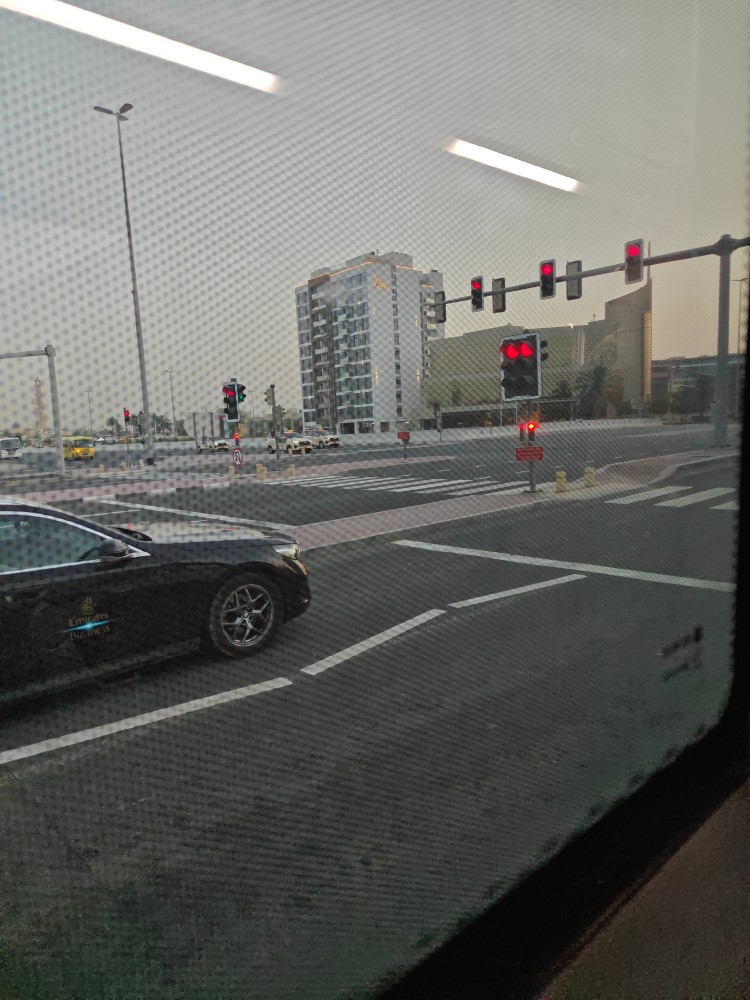 This image captures a view through a window, likely from a vehicle, showcasing a street scene in Dubai, United Arab Emirates. The foreground features the side of a black car with the "Emirates Business" logo visible on its door. The car is positioned on a multi-lane road with white lane markings and pedestrian crosswalks.
In the middle ground, a traffic intersection is visible with multiple traffic lights. Several of these traffic lights are illuminated with red signals, indicating that traffic should stop. One prominent traffic light pole has a row of four traffic lights, all showing red. Another traffic light closer to the viewer displays two red lights.
Beyond the intersection, various buildings are present. A modern, multi-story building with many windows and balconies stands to the left. To the right, a taller building with a lighter facade is visible, along with other structures that appear to be part of the urban landscape. Palm trees are interspersed among the buildings, suggesting a warm climate.
In the background, the sky is overcast, giving the scene a muted, diffused light. The overall impression is of a typical urban environment in Dubai, with active road infrastructure and modern architecture. The image is slightly blurred due to the window screen.