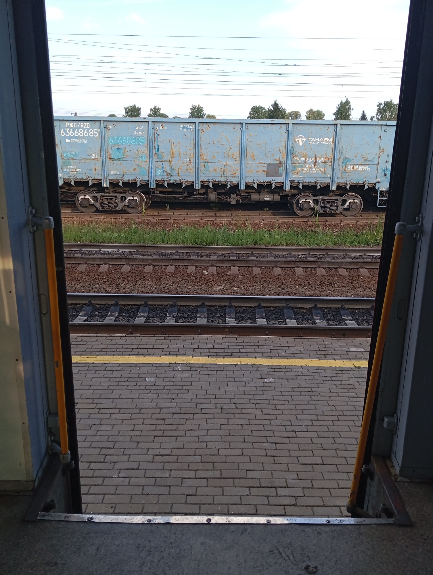 This image depicts a view from the doorway of a train carriage looking outwards. The primary subject is a blue freight train carriage, showing its side with visible markings and a set of wheels. The train is situated on railway tracks, with gravel ballast and some grass visible along the side. Adjacent to the tracks is a paved area, likely a platform, marked with a yellow line. The sky is bright and appears to be daytime, with some scattered clouds, suggesting fair weather.
The visible text on the train carriage includes Cyrillic lettering and numbers, indicating the origin, type, and manufacturing details of the wagon. Specifically, "РЖД/RZD 63668685" appears prominently, along with "ТАHДЕМ" and manufacturing dates such as "13.06.2018" and "13.06.29". The overall scene is one of a functional, operational railway environment. There are no people visible in the frame. The perspective suggests the photograph was taken from a stationary train or a train that has just stopped at a station. The composition, framed by the dark interior of the train doorway, draws attention to the train and its surroundings.