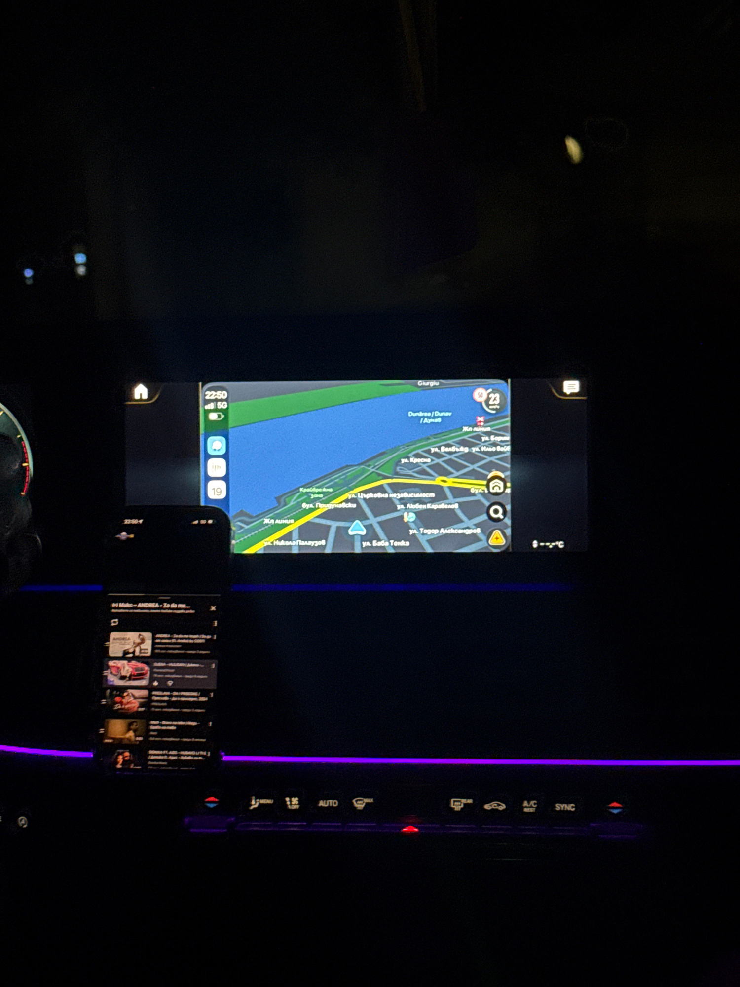 The image shows a car's dashboard at night, with a navigation screen displaying a map of the city of Ruse, Bulgaria. The navigation screen shows the Danube River on the left side, with a road running along its bank. Several street names in Cyrillic script are visible on the map, including "ул. Басараби" (ul. Basarabi), "бул. Приморски" (bul. Primorski), and "ул. Георги Димитров" (ul. Georgi Dimitrov). The map also shows various points of interest, such as a train station and a museum.
Below the navigation screen, a smartphone is visible, displaying a playlist of songs. The car's interior is illuminated by ambient purple lighting. The overall scene suggests a nighttime drive through the city of Ruse.