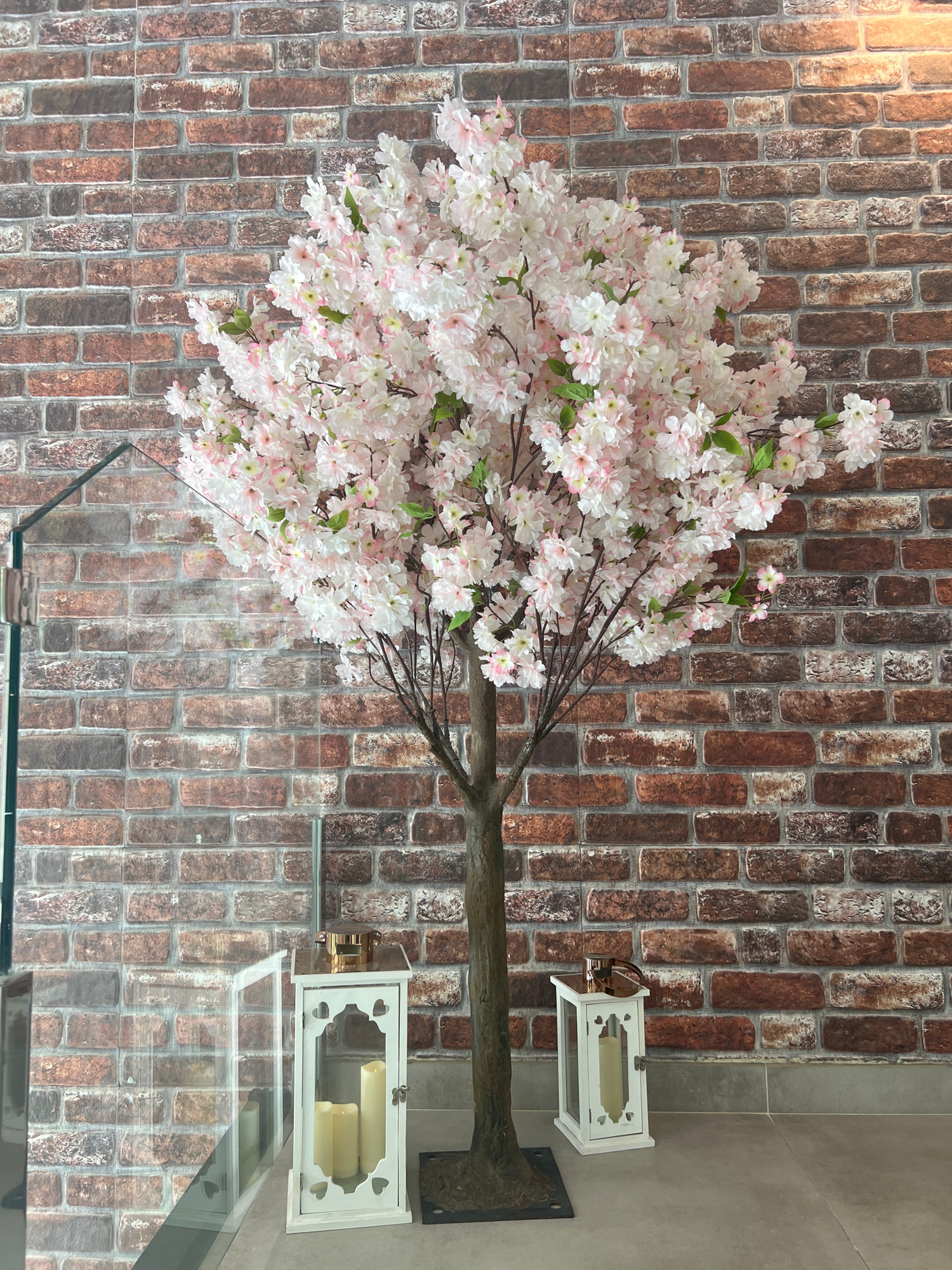 This is an indoor shot against a brick-patterned wall. A large, artificial cherry blossom tree with pink and white flowers dominates the left side of the frame. The tree trunk is brown and textured. Two white, decorative lanterns with glass panes are placed on a glass surface to the right of the tree. Each lantern contains cream-colored candles. The lighting suggests it is daytime, and the setting appears to be a commercial or residential interior, possibly a shop or a home with a decorative display. There is no visible text in the image.