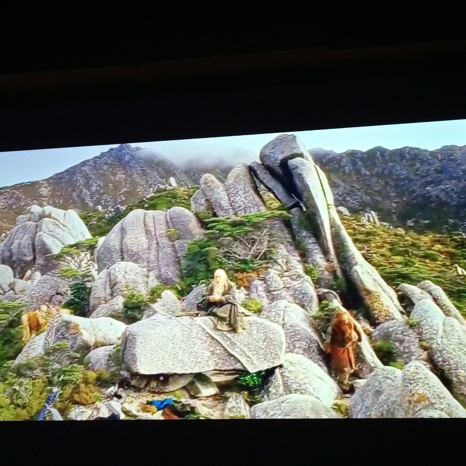 This image depicts a rugged, mountainous landscape with scattered, large grey rocks and patches of green vegetation. In the foreground, two figures are visible. The first figure, on the left, is seated on a flat rock, wearing a long, grey robe and has a long white beard. The second figure, on the right, is standing and appears to be wearing brown and orange clothing. The background shows more rocky terrain and distant mountains shrouded in mist. There is no indication of the city of Zam'yany, Russia in this image.