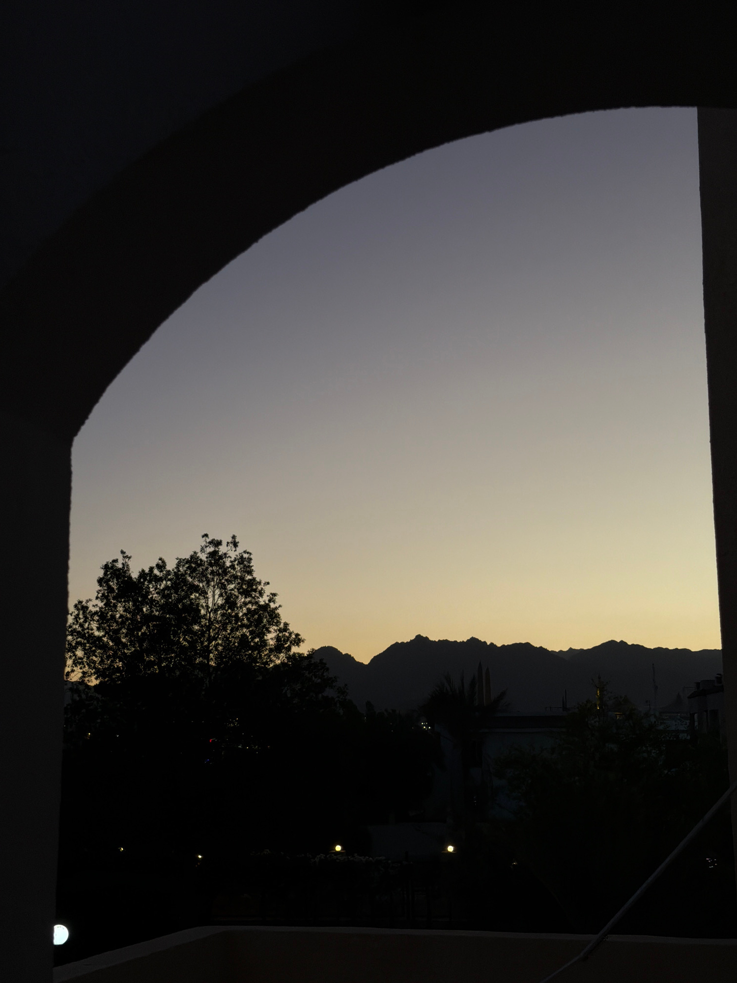 The image captures a twilight scene through an arched opening, framing a silhouette of mountainous terrain in the distance. The sky transitions from a deep twilight blue at the top to a soft yellow near the horizon. In the foreground, silhouetted trees obscure much of the landscape, with a prominent, leafy tree on the left and a cluster of shorter trees and buildings to the right. Several faint lights are visible amongst the trees, suggesting artificial illumination within a populated area. The overall impression is of a tranquil evening, likely in a suburban or semi-rural setting, with the natural beauty of the mountains providing a dramatic backdrop. The presence of buildings and lights hints at the proximity of a settlement, consistent with the environment of Sharm El Sheikh, Egypt.
