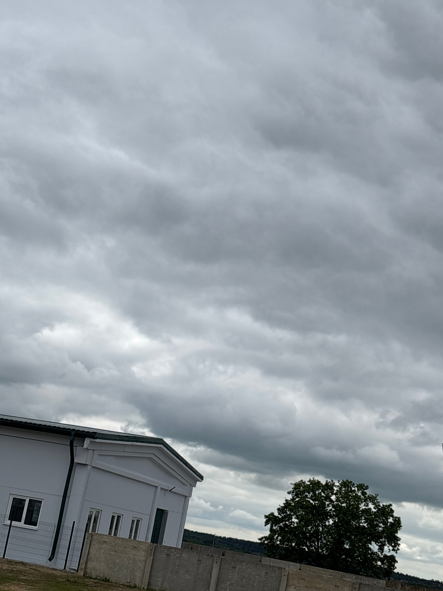 This media file depicts a building with a grey, segmented exterior, likely a wall or a series of connected structures, on the right side of the frame. The building has several windows and a dark-colored roofline with visible gutters. A fenced area is also apparent in front of the building. To the left of the building, a large, dark green tree with full foliage stands out against a backdrop of dramatic, overcast skies. The sky is filled with heavy, grey clouds, suggesting a cloudy or potentially stormy day. The overall scene is viewed from a low angle, looking upwards towards the sky and the upper portions of the building and tree. The image appears to be taken outdoors, with the location context suggesting it is in the Kapylsky Selsovet, Belarus. There are no people or discernible activity taking place in the image, and no visible text. The time of day is not precisely indicated, but the lighting suggests it could be daytime with overcast conditions. The emotion evoked by the image is one of somberness or anticipation, due to the heavy clouds and the starkness of the architectural elements.