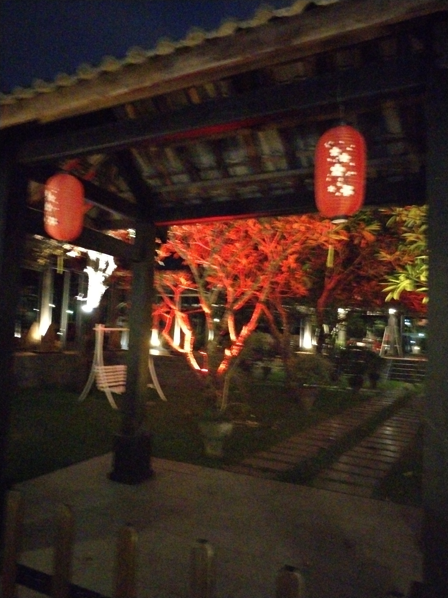 The image depicts an outdoor courtyard area at night. Two red paper lanterns with intricate white patterns are visible, one hanging from a wooden structure on the left and another suspended higher up. The courtyard features trees illuminated with red lights, creating a vibrant, warm glow. A white wooden swing set is present in the mid-ground. A wooden pathway winds through the courtyard. The scene is framed by a dark, wooden roof structure. The location is identified as a courtyard in Bac Nha Trang Ward, Vietnam.