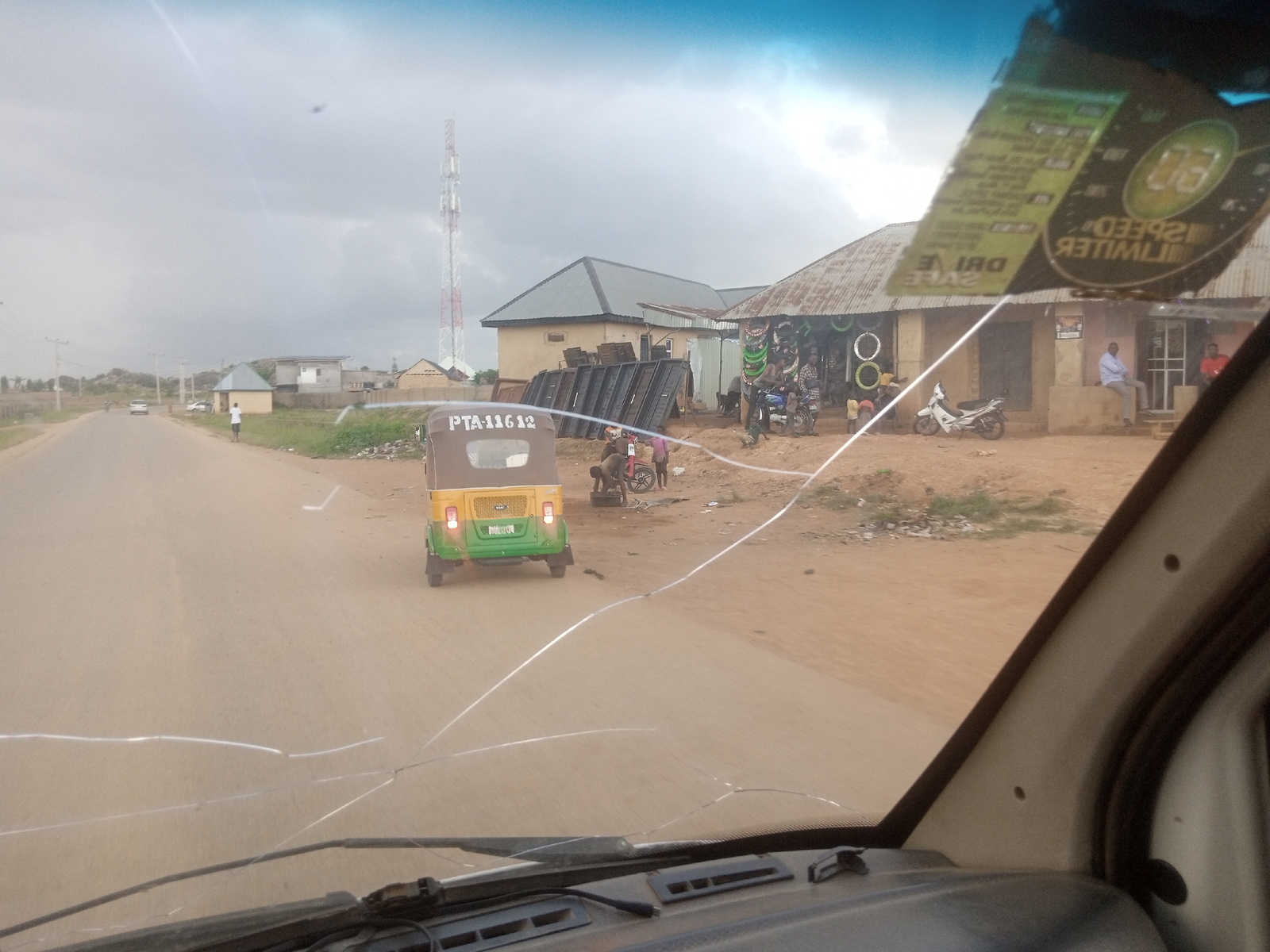 This media file captures a scene from inside a vehicle, looking out through a cracked windshield onto a dusty road. The foreground shows the dashboard of the vehicle and the cracked windshield itself, which distorts the view slightly.
The main subjects in the scene are a bright yellow and green auto-rickshaw (also known as a tuk-tuk) driving away from the camera on the right side of the road. The license plate on the back of the auto-rickshaw is visible and reads "PTA 116 12". Further ahead, several people are gathered around what appears to be a shop or stall selling metal items, possibly for construction or repair, stacked in long, dark pieces. There are also tires displayed outside the shop. A white motorcycle is parked in front of the shop.
In the background, a tall radio tower with red and white sections stands prominently against a cloudy sky. There are several buildings visible, some with corrugated metal roofs, suggesting a somewhat rural or developing area. The ground is a light brown, dusty earth. The lighting suggests it is daytime, but the overcast sky indicates potentially cloudy or impending weather. The overall impression is of a normal day with ongoing activity in a roadside community. A sticker with text is affixed to the inside of the windshield, partially obscuring the view. Some text is readable on the sticker, including "SPEED LIMITER" and "DRIVE SAFE".