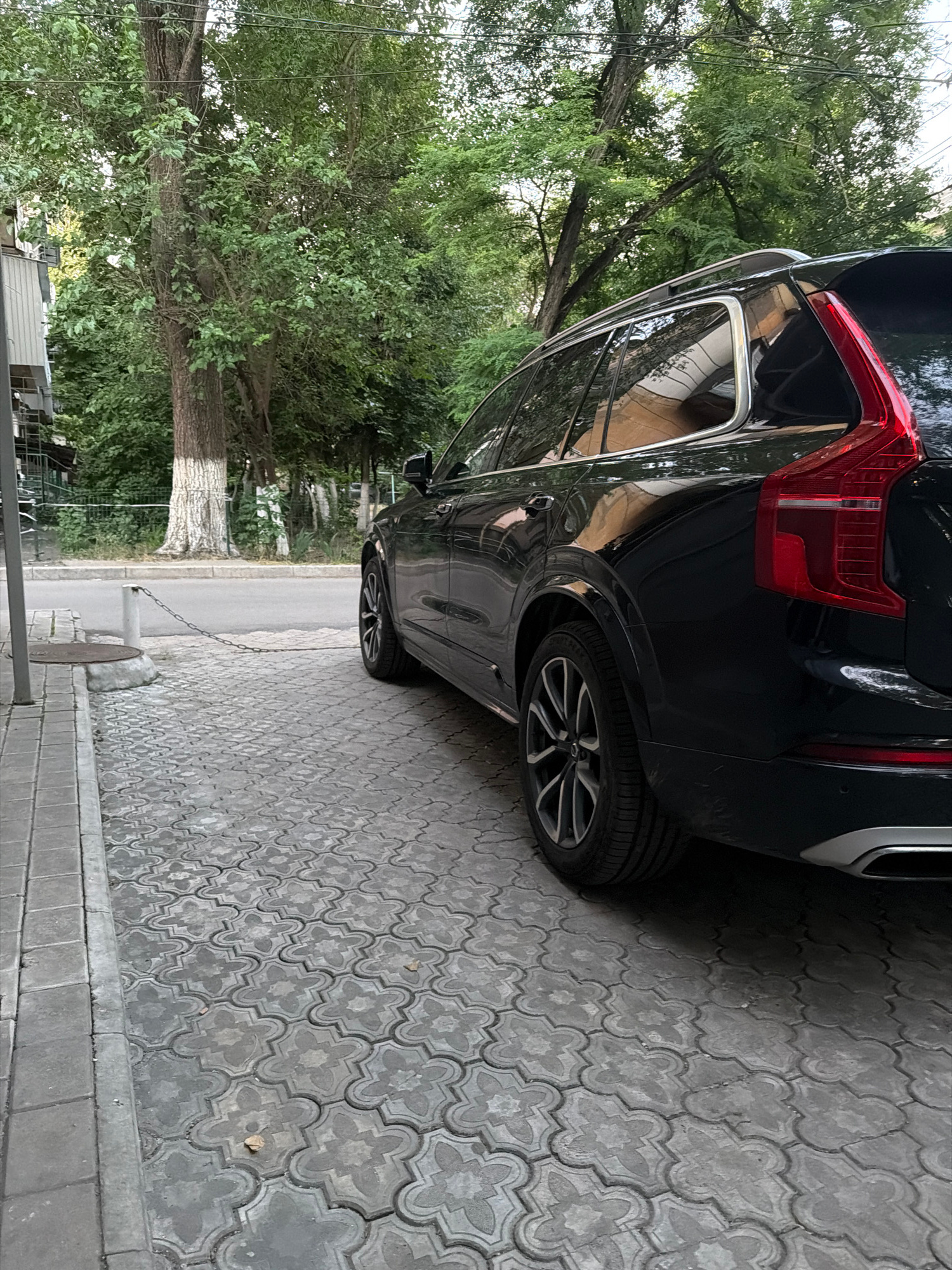 A black Volvo SUV is parked on a patterned gray paved surface next to a sidewalk. A gray concrete curb separates the pavement from a street. A metal chain connects a short concrete post on the curb to a manhole cover. Trees with green foliage line the street and extend into the background. A building with a white-trimmed facade is partially visible on the left. The sky is overcast.