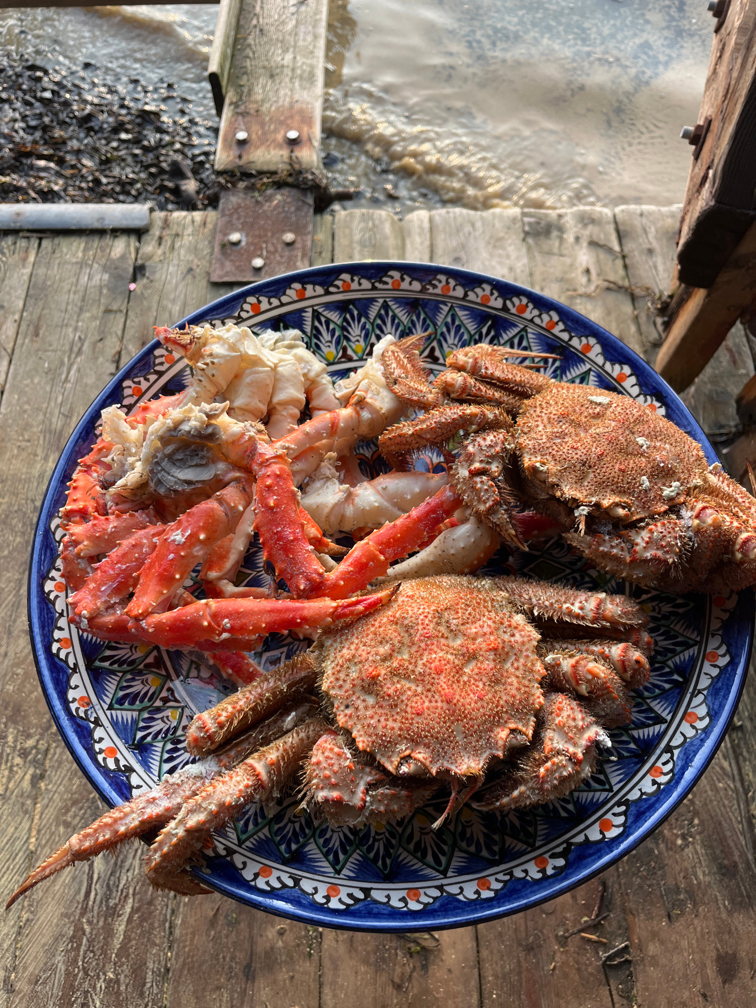 A close-up, high-angle shot captures a blue and white patterned ceramic plate filled with cooked crabs and king crab legs. The crabs are reddish-orange and hairy, with some white bits of flesh showing. The king crab legs are larger and brighter red, with some white exposed meat at the joints. The plate rests on a rustic wooden surface, possibly a pier or deck, with a glimpse of dark water and pebbles in the background. The water appears choppy, suggesting a breezy day. The scene is likely outdoors, possibly in a coastal area like Sovetskaya Gavan, Russia, given the presence of seafood and the weathered wooden structures. There are no people visible in the image.