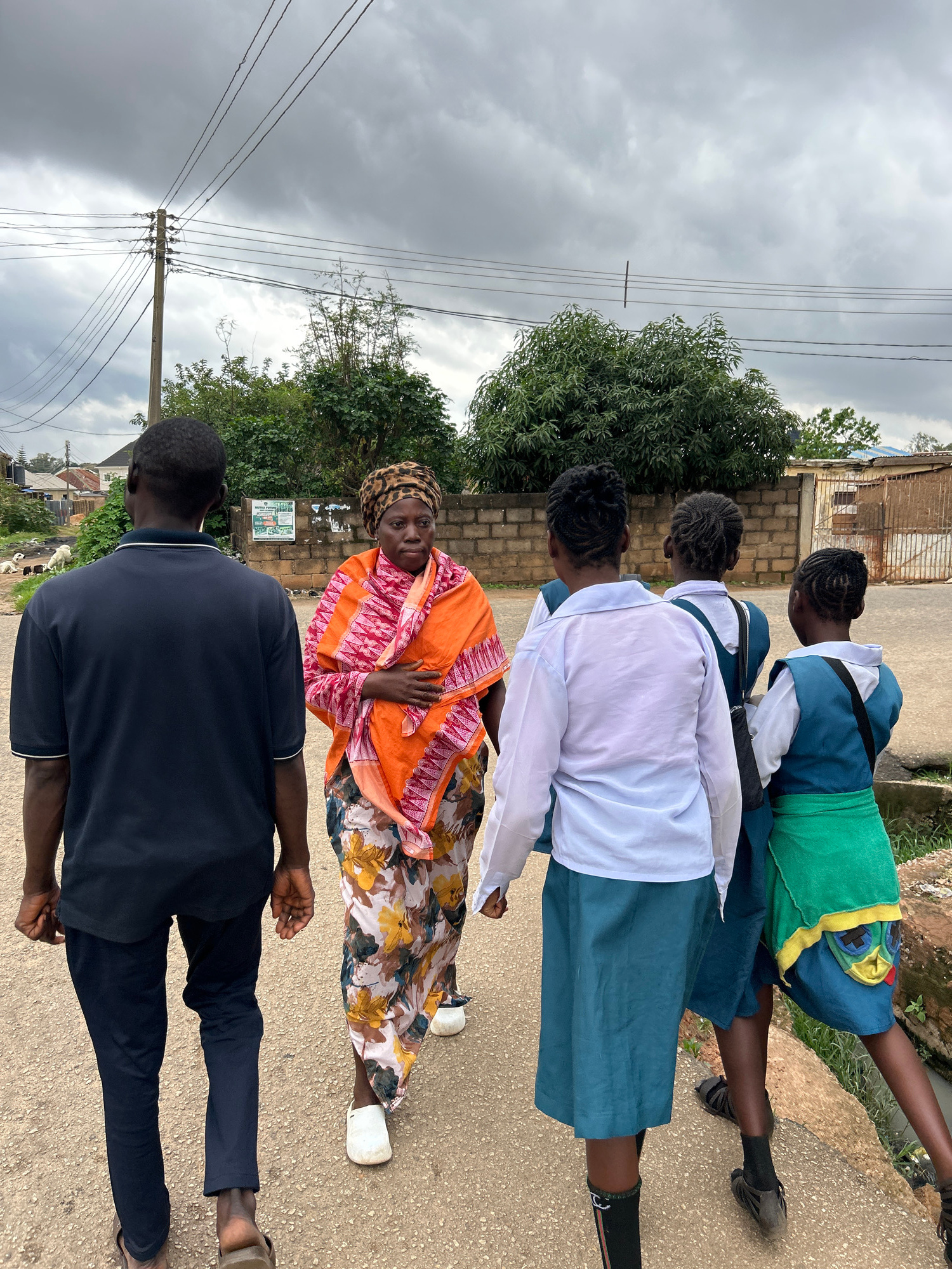 In Jos, Nigeria, under a cloudy sky, a group of five individuals walks away from the camera on a paved road. The foremost person, a man wearing a navy blue polo shirt and dark trousers, walks with his back to the viewer. Beside him, a woman in a vibrant orange and pink patterned wrap and a floral skirt walks with her left hand across her chest. Behind them, three young girls in school uniforms walk together. The two girls in the middle and on the right wear teal jumpers over white shirts, with the girl on the right also wearing a green wrap around her waist. The girl on the left wears a white shirt and a teal skirt. The environment includes a brick wall to the right, some trees in the background, and a utility pole with wires overhead. In the distance on the left, houses and more trees are visible, with a few goats grazing in a grassy area.