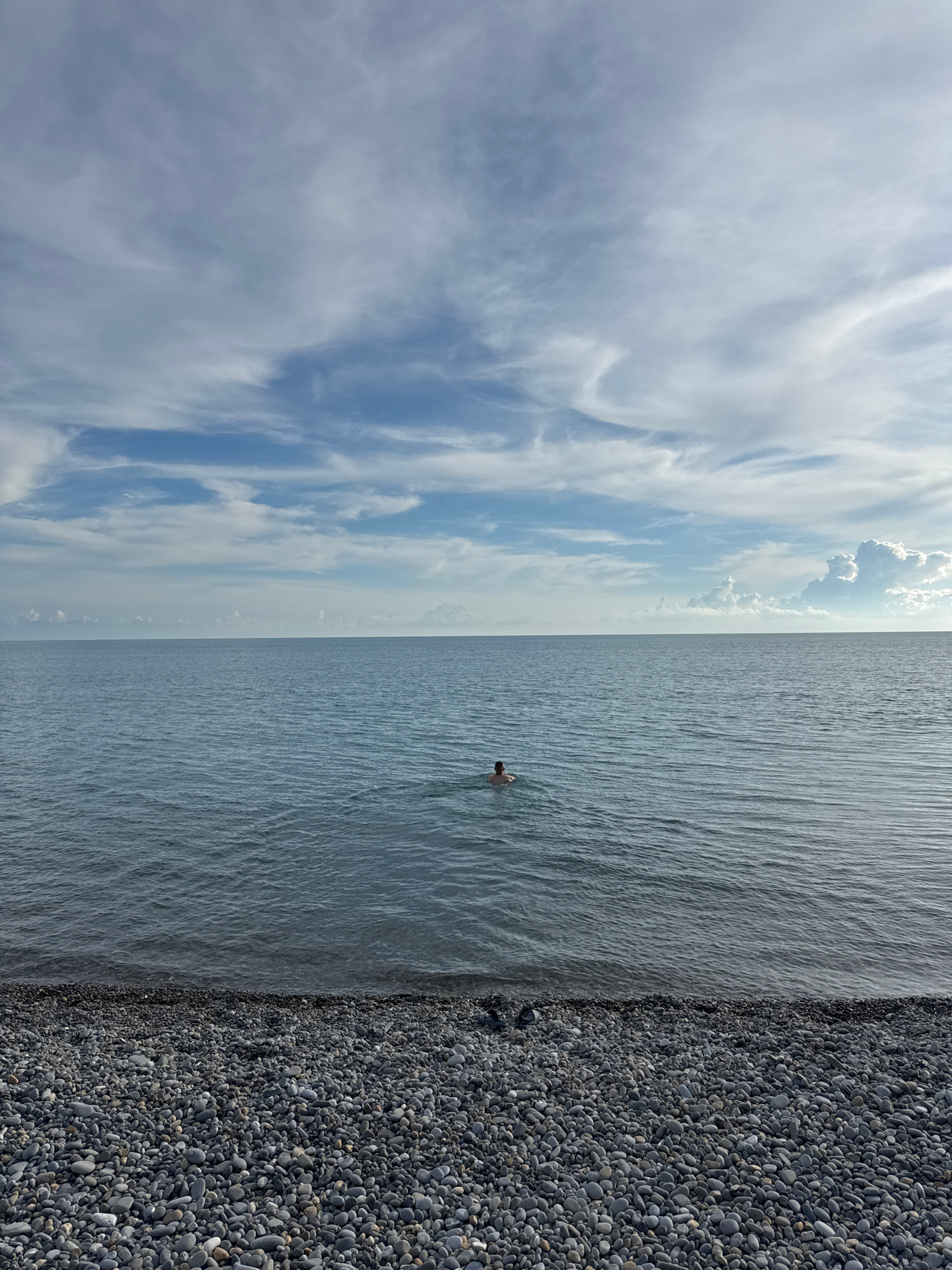 A person is swimming in the calm, rippling waters of the Black Sea, seen from a pebble beach. The sky above is a vast expanse of blue with scattered, wispy white clouds. The horizon is sharply defined where the water meets the sky. The foreground is dominated by the dark, rounded stones of the beach. The scene suggests a peaceful moment by the sea in Sochi, Russia.