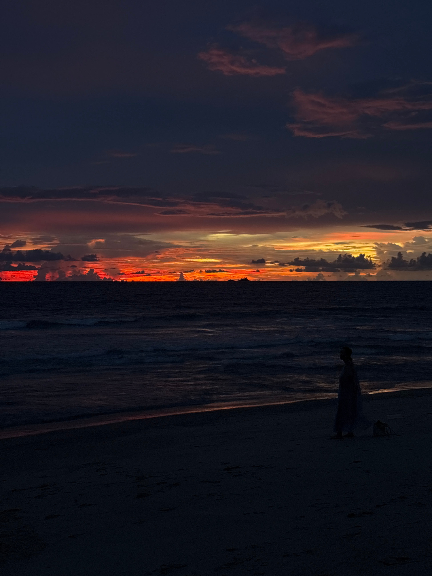 This media file captures a dramatic sunset scene on a beach. The image is oriented vertically, with the left side dominated by a dark sky filled with dramatic clouds tinged with hues of orange, red, and yellow from the setting sun. The right side of the image features a dark, textured surface that appears to be sand or a dark, rough terrain. Between the sky and the dark foreground, a band of intensely colored light from the sunset is visible, reflecting on what seems to be water or wet sand.
In the upper right portion of the image, a small, silhouetted figure stands on the dark surface, facing towards the sunset. The figure appears to be wearing a light-colored garment, possibly a dress, and their posture is still, suggesting they are observing the sunset. The overall mood evoked by the image is one of awe and tranquility, with the vibrant colors of the sunset contrasting with the dark, quiet surroundings. The time of day is clearly sunset, and the weather appears calm, with no strong winds evident. Location cues are not explicit, but the provided context suggests a beach in Choeng Thale, Thailand. There is no visible text in the image.
