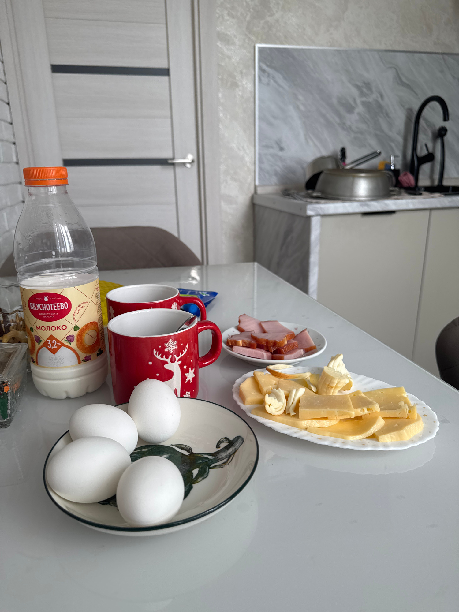 An indoor scene, likely a kitchen or dining area, features a white table with various food items. In the foreground, a plate holds four white eggs, one of which is partially obscured. To the right of the eggs, a white oval plate is arranged with numerous slices of yellow cheese, some of which are cut into decorative shapes. Behind the cheese plate, a small white round plate contains several slices of pinkish ham. A red mug with a snowflake and reindeer design, containing a spoon, sits next to another identical red mug. In the background, a clear plastic bottle of milk with an orange cap is positioned behind the mugs. A portion of a kitchen counter is visible with a faucet and some cookware in the background, suggesting a domestic setting. The overall lighting is bright, indicating an interior space.