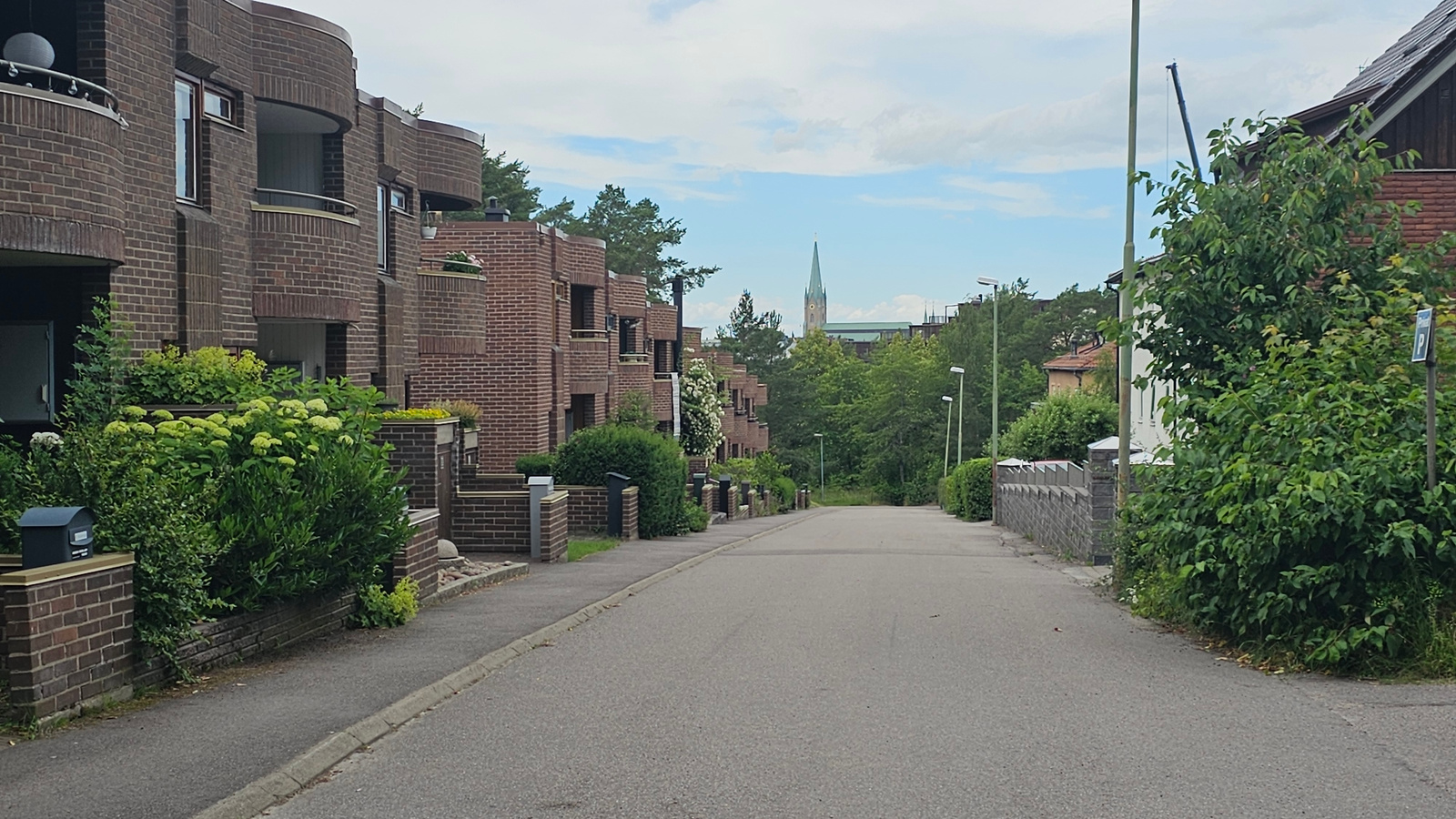 A low-angle, eye-level shot captures a residential street scene in Linköping, Sweden. The street slopes gently upwards and recedes into the distance, lined on the left by multi-story brick buildings with balconies and lush green landscaping. On the right, the street is bordered by a low stone wall, more trees, and the side of a building.
The architecture of the brick buildings on the left is characterized by rounded corners and a modernist aesthetic. Planters with vibrant green foliage and white flowers are visible on balconies and along the front of the buildings. Mailboxes are mounted on brick pedestals.
In the background, a church steeple with a green roof pierces the sky, surrounded by a canopy of dense green trees. The sky is partly cloudy, suggesting a pleasant day. The overall impression is one of a quiet, well-maintained suburban neighborhood. No people are visible in the frame, and no specific activity or event is taking place. The time of day appears to be daytime, given the lighting conditions. The location is identified as Linköping, Sweden. There is a blue parking sign visible on the right side of the image with a white letter "P".