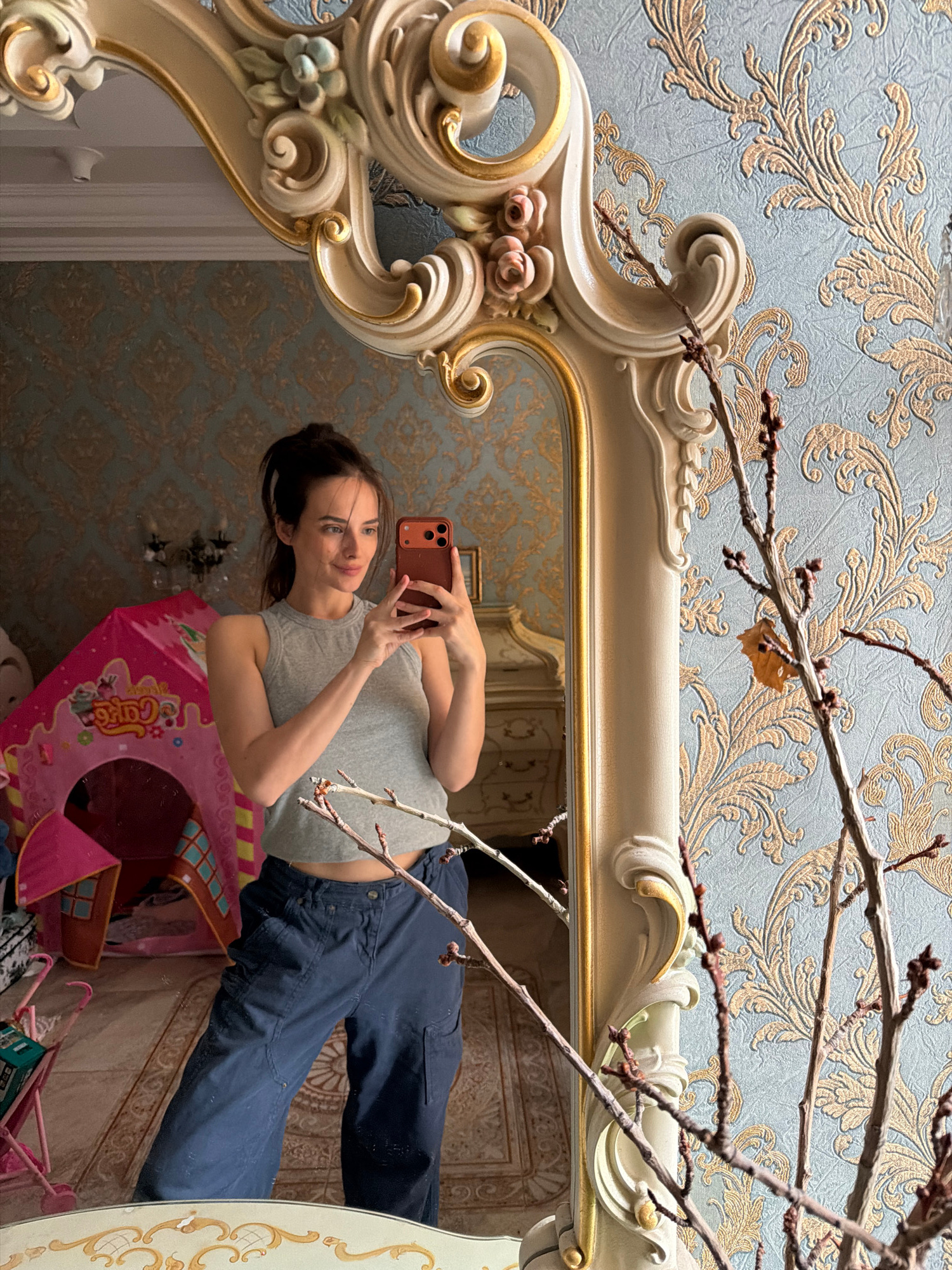A woman with dark hair pulled into a ponytail is taking a mirror selfie in what appears to be a bedroom. She is wearing a grey tank top and navy blue cargo pants. The mirror is ornate and framed with gold accents and decorative carvings. In the reflection, behind her, there is a pink children's play tent and a pink toy stroller. The wallpaper in the room has a light blue background with a gold damask pattern. In the foreground of the mirror, branches with small buds and a single dried leaf are visible. There is no indication of the specific location in Ufa, Russia.