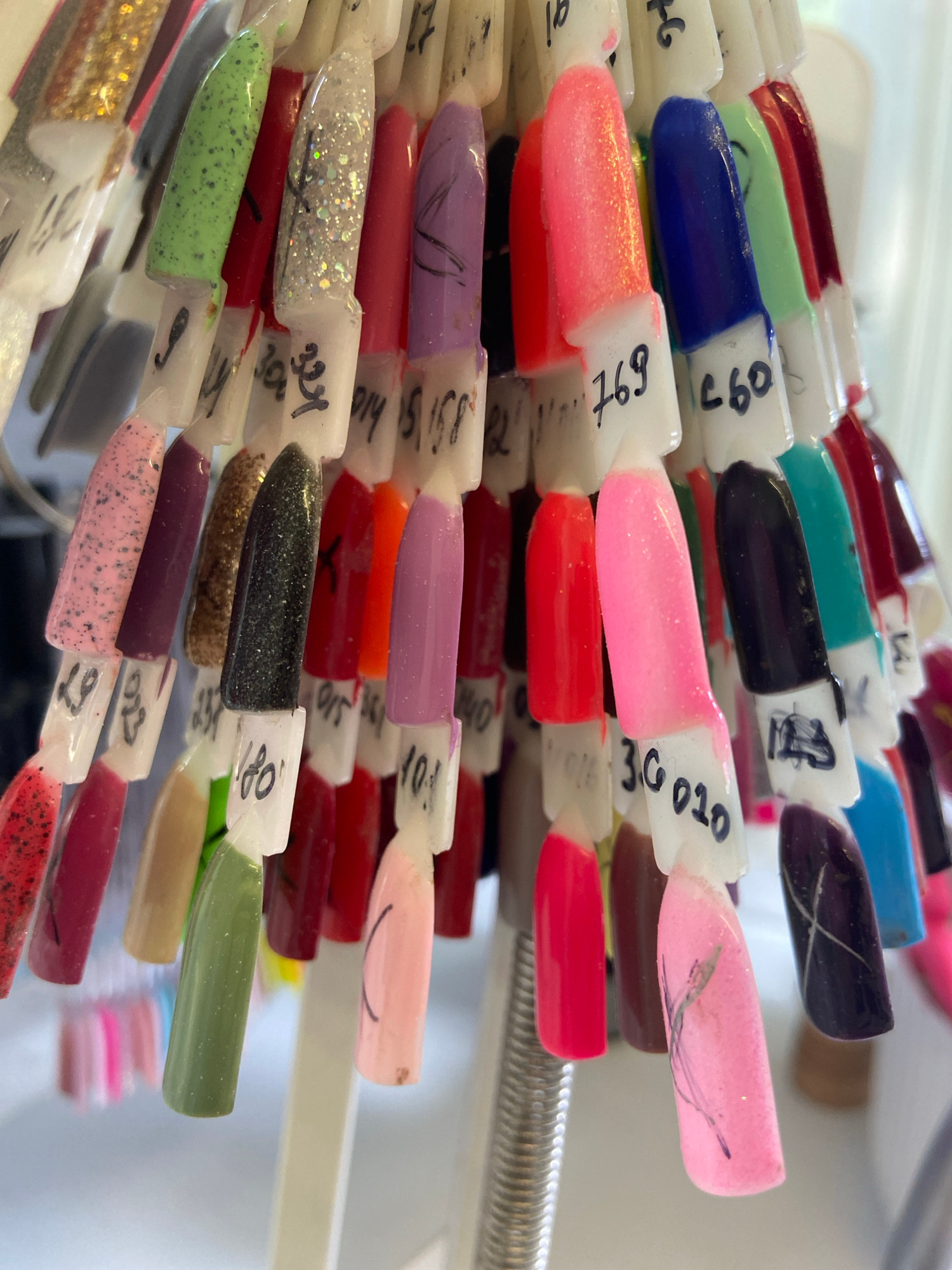 A close-up, eye-level, medium shot captures a display rack filled with numerous colorful nail polish swatches. The swatches, made of plastic, are shaped like elongated teardrops and are attached to white plastic handles. Each handle has a handwritten number or code on it, likely for identification. The nail polish colors vary widely, including glittery gold, speckled green, shimmery silver, vibrant purple, neon pink, deep blue, black, and various shades of red, pink, and brown. Some swatches feature subtle embellishments, like glitter or delicate line art on a pink swatch. The swatches are arranged in multiple rows, some overlapping, creating a dense, textured display. The lighting is bright, suggesting an indoor environment, possibly a salon or beauty supply store. The background is blurred and out of focus, but hints of a light-colored wall or partition are visible. The image focuses on the detail and variety of the nail polish colors.
