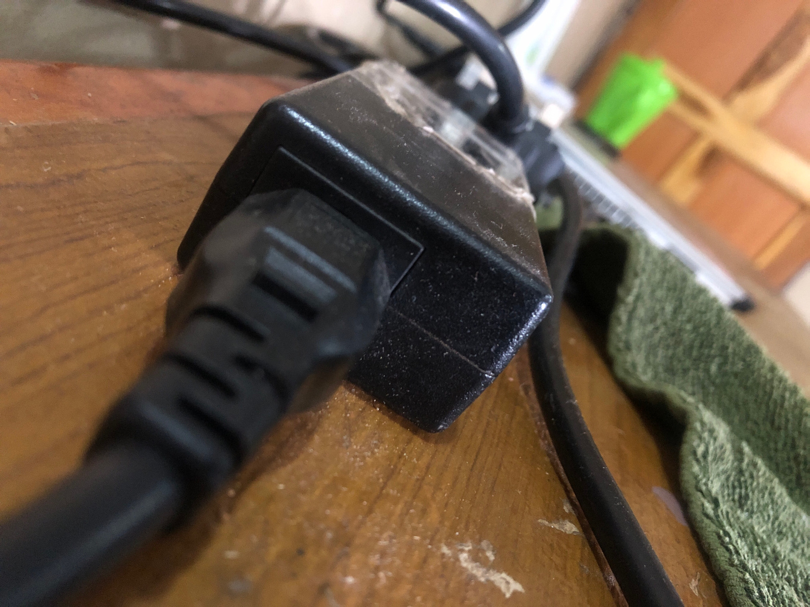 A close-up, low-angle shot shows a black power adapter with a plug inserted into it, lying on a wooden surface. The adapter and its plug are covered in a light layer of dust. To the right of the adapter, a green, textured fabric is visible, with a black cable running along the bottom of the frame. In the background, a blurred green object and wooden structures are visible. The scene appears to be indoors, possibly on a desk or table.