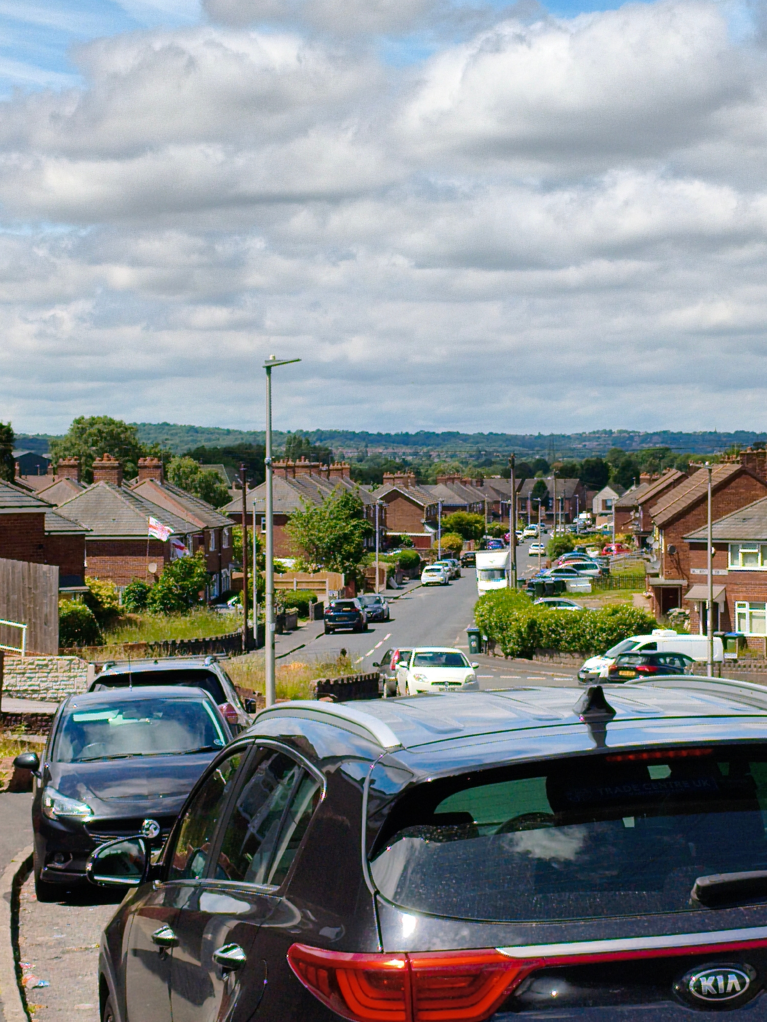 This is a photograph taken from a slightly elevated position, looking down a residential street lined with red-brick houses and parked cars. The sky is filled with large, fluffy white clouds against a blue background. The street is asphalt and appears to be a residential area in Sandwell, United Kingdom.
In the foreground, the rear of a dark-colored SUV, possibly a Kia, is prominently displayed, facing away from the camera. To the left of this SUV, a dark-colored hatchback is parked, facing the camera, with its headlights on. Further down the street, more cars are visible, including a white hatchback, a white van, and other vehicles. The houses have pitched tile roofs and visible chimneys. There are also trees and green foliage along the street and in front of the houses. A lamppost stands on the right side of the street. The overall scene depicts a typical suburban street on a cloudy but bright day.