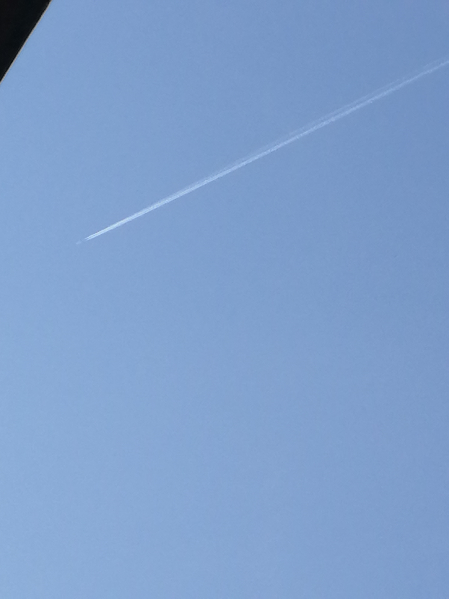 A clear, bright sky is the setting for this image. The sky is a light blue color, suggesting it is daytime, possibly late morning or early afternoon, with good visibility. There are no clouds visible, indicating fair weather. A faint white contrail, typical of an airplane flying at high altitude, stretches diagonally across the upper portion of the frame from the bottom left to the top right. The contrail is thin and wispy, suggesting the airplane is some distance away. A dark, angular object is visible in the very top left corner of the image, partially obscuring the sky. This object could be part of a building, a tree, or another man-made structure. There is no text visible in the image.
