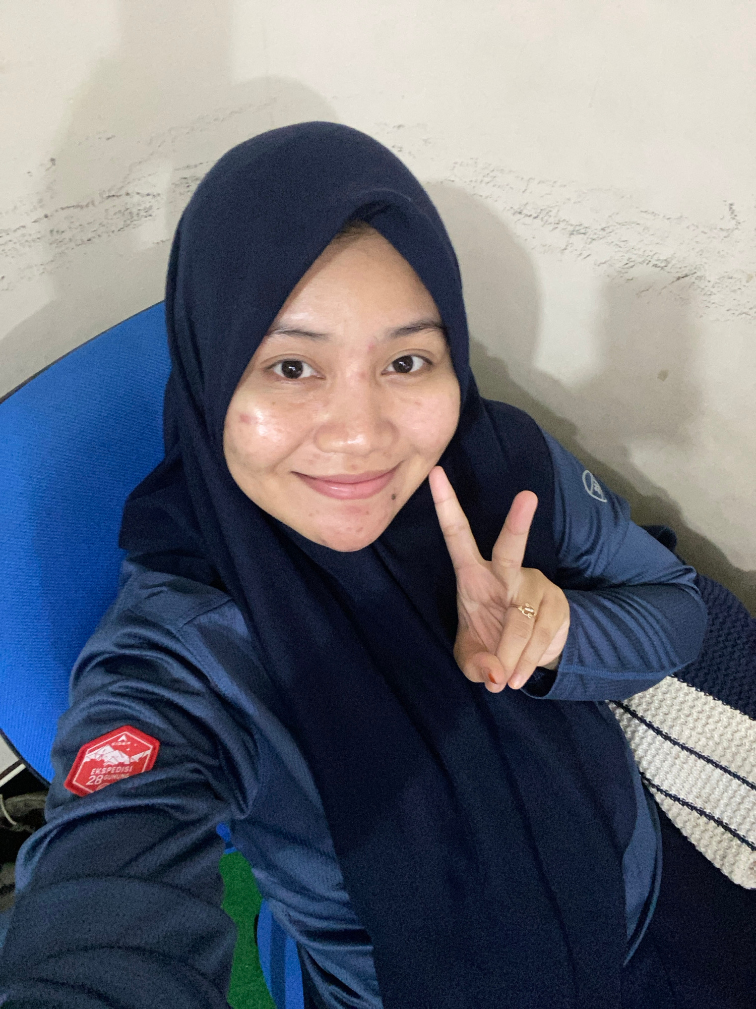 A young woman wearing a dark blue hijab and a dark blue long-sleeved shirt is lying down and taking a selfie. She is smiling and holding up two fingers in a peace sign. A ring with the letter "A" is visible on her right ring finger. The woman has fair skin and some acne on her face. She is lying on a blue mat, and in the background, there is a textured wall and a blanket with white and dark blue stripes. A patch with text and a graphic is visible on her left sleeve. The scene appears to be indoors. The text on the patch reads "EKSPEDISI 28".