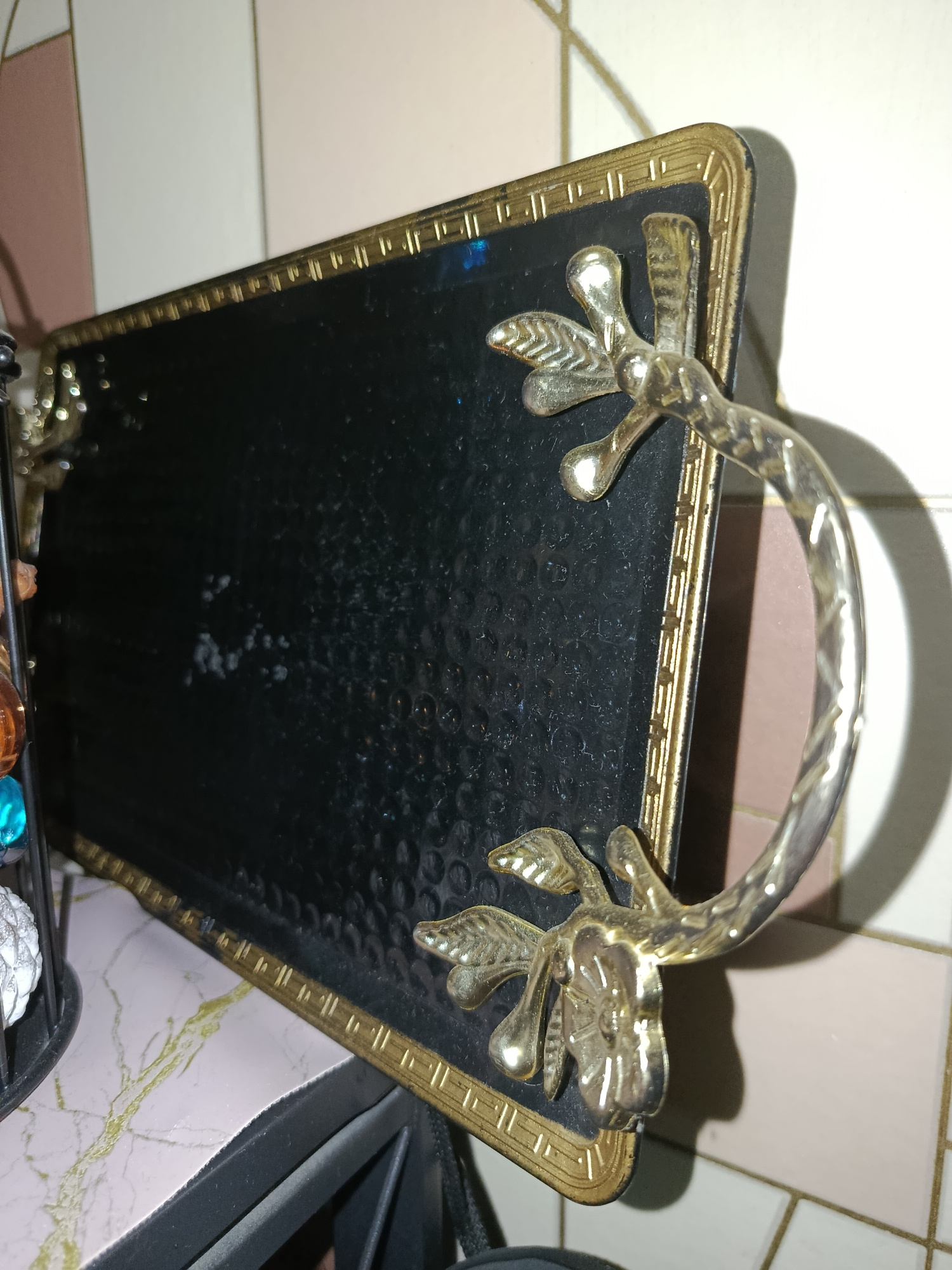 An ornate, rectangular tray with a black, textured surface and a gold-colored, decorative border is positioned on a marble-patterned countertop. The tray features two handles shaped like stylized branches with floral embellishments, made of a metallic material with a gold finish. The background consists of a tiled wall with alternating pink and white square tiles. On the left side of the tray, a black metal stand holds decorative items, including colorful beads. The scene appears to be indoors, likely in a domestic setting, with artificial lighting casting shadows. The image was taken in Tashkent, Uzbekistan.