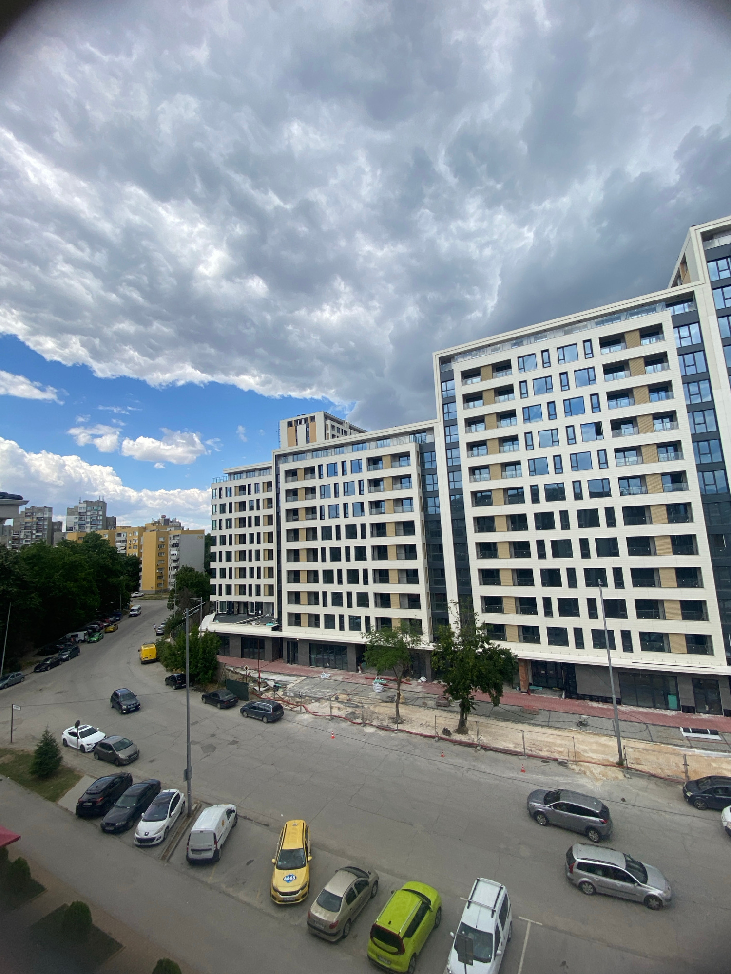 This media file captures an urban scene from an elevated perspective, likely a balcony or window. The primary subjects are modern apartment buildings and a street lined with parked cars. The buildings are tall and multi-storied, with numerous windows, suggesting a densely populated residential or mixed-use area.
The street below shows a typical urban setting with asphalt paving and visible parking spaces. A variety of cars are parked along the street, including sedans and hatchbacks in different colors like white, gray, black, and yellow. Some of the cars appear to be taxis, identifiable by their bright yellow color and potential markings, though any text on them is not clearly legible. There are also trees planted along the street and sidewalks, adding a touch of greenery to the urban landscape.
The sky is a dominant element, filled with dramatic, swirling gray clouds, interspersed with patches of bright blue sky. This suggests a dynamic weather pattern, possibly a mix of overcast conditions and sunshine. The lighting implies it is daytime, but the cloudy sky may be softening the sunlight, creating a diffused illumination.
The overall impression is a snapshot of everyday life in a European city, specifically Plovdiv, Bulgaria, as indicated by the location context. There are no visible people in the scene, and the focus is on the architecture and the vehicles. The composition, with the buildings and street tilted, suggests the photo was taken at an angle, possibly looking down and to the side. The scene is devoid of any apparent emotional cues, presenting a factual representation of the urban environment.