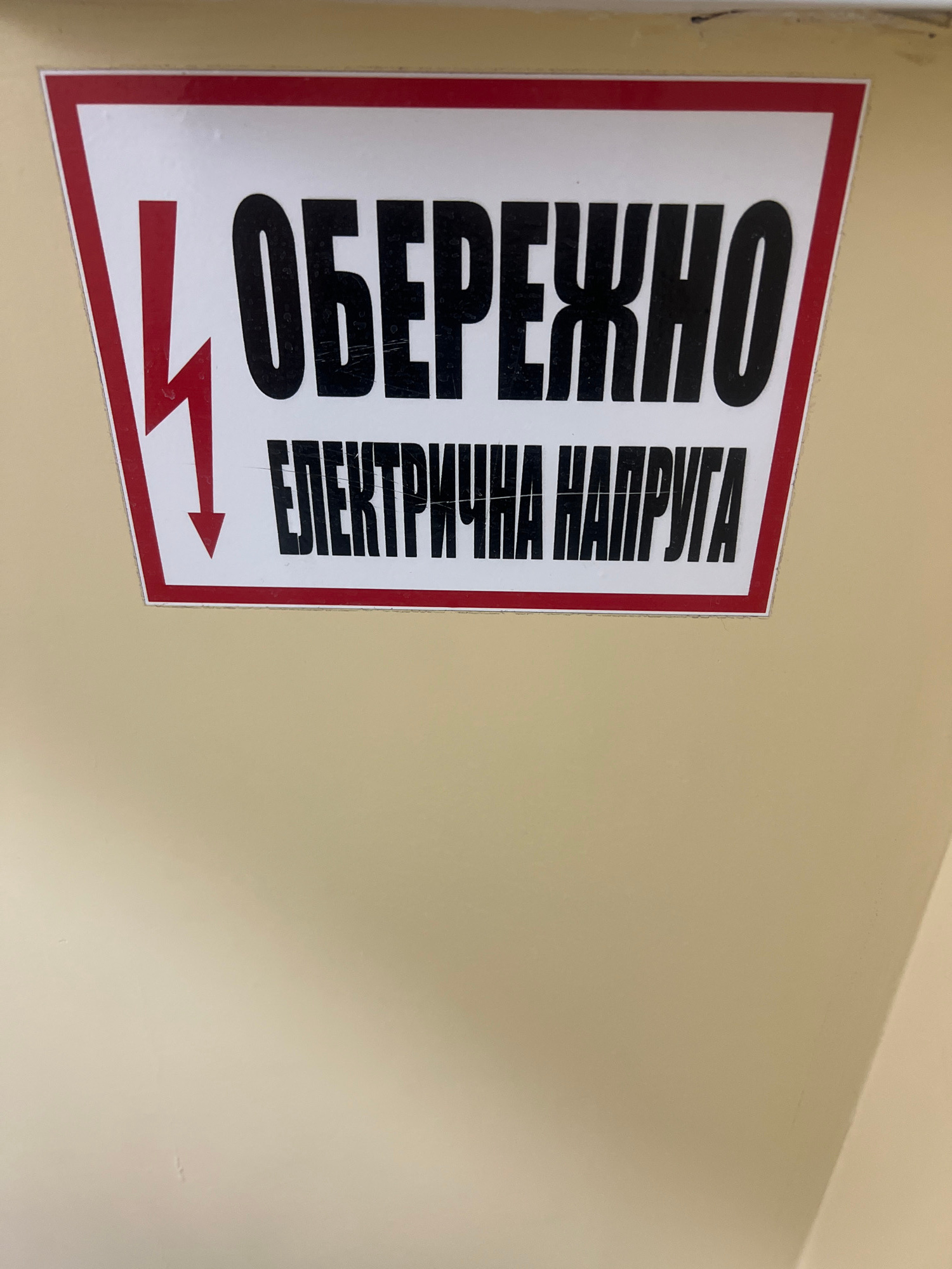 The image shows a white rectangular sign with a red border, mounted on a light beige wall. The sign features a red lightning bolt symbol on the left, pointing downwards, followed by black text. The top line of text reads "ОБЕРЕЖНО" (OBEREZHNO), meaning "Caution" or "Beware." The second line reads "ЕЛЕКТРИЧНА НАПРУГА" (ELEKTRICHNA NAPRUHA), meaning "Electric Voltage." The sign appears to be an electrical hazard warning. The location in Kropyvnytskyi, Ukraine is not specified in the image itself.
