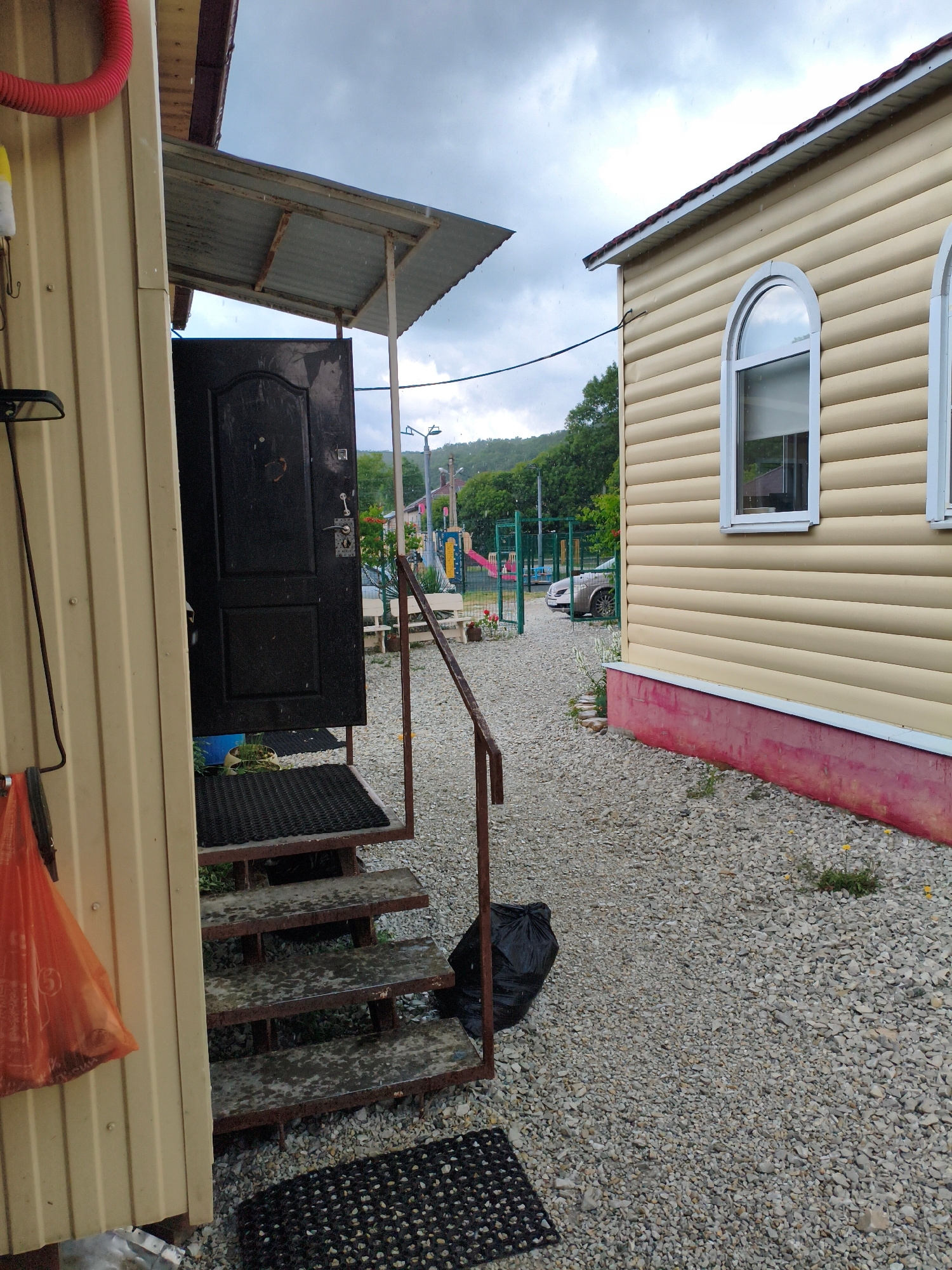 The image depicts an exterior view of a building with light-colored, horizontally ribbed siding and a dark-colored door with an open entryway leading to a set of metal stairs. The stairs have black rubber mats on the top two steps and appear wet. A red hose is visible coiled on the left side of the building, and an orange plastic bag hangs from a hook. The ground is covered in gravel. To the right, another building with similar siding and arched windows is visible, with a red-painted foundation. In the background, a playground area with colorful equipment and a green fence can be seen. A car is parked near the playground. The sky is overcast with visible raindrops. The scene appears to be in a residential or small business area, possibly in the city of Vozrozhdeniye, Russia, given the context provided.