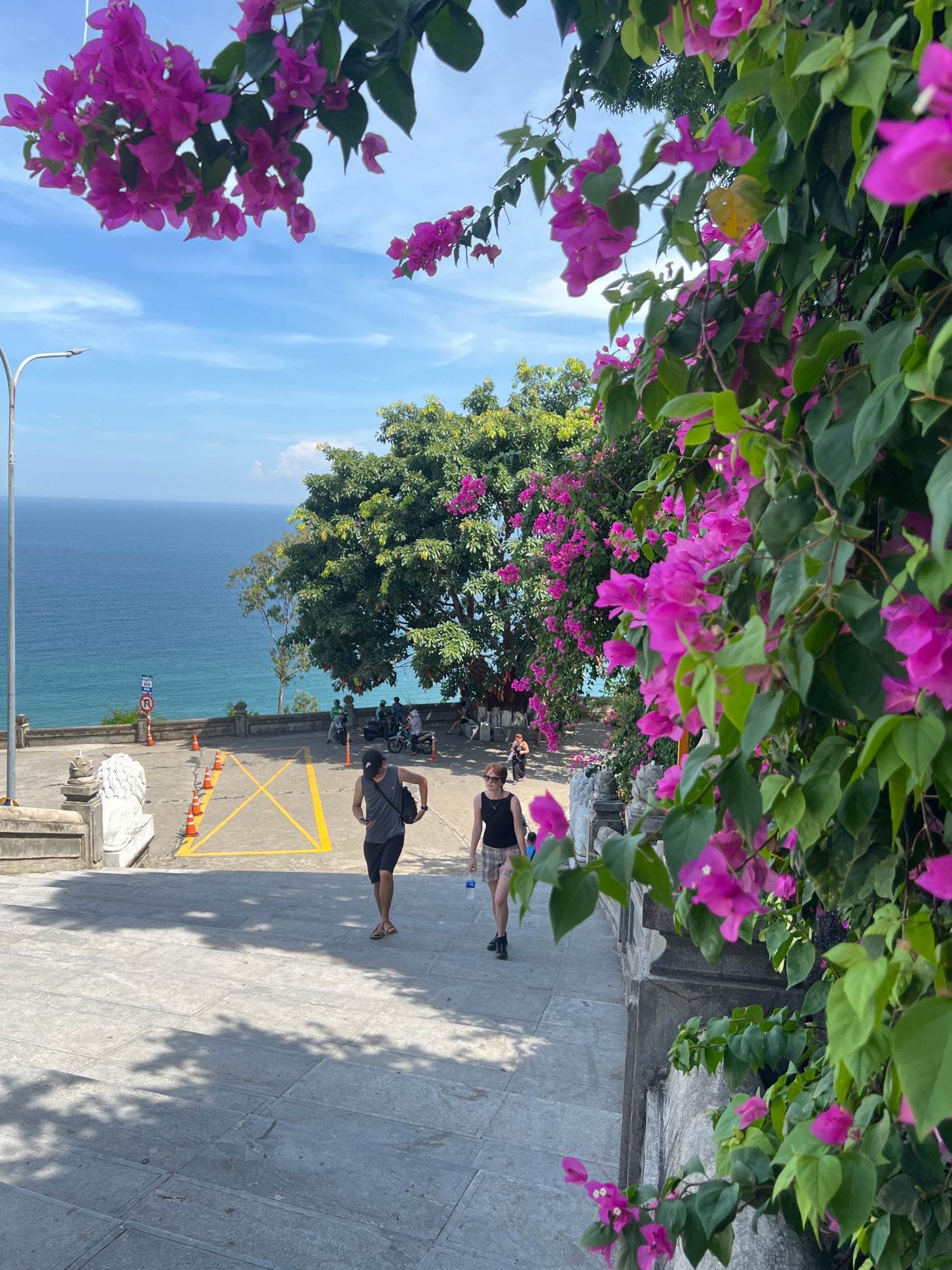 A wide-angle, eye-level shot captures a view from a paved staircase looking down towards the ocean and a parking area. In the foreground, vibrant pink bougainvillea flowers cascade down a wall on the right side of the frame, with lush green leaves and branches partially obscuring the view. Two people are walking down the staircase in the mid-ground: one individual in a dark grey sleeveless shirt and black shorts with a black bag slung over their shoulder, and another person in a black tank top and plaid shorts. Further down, a group of people are gathered near parked motorbikes in a paved area marked with yellow lines, with traffic cones lining the side. The background features a clear blue sky with some wispy clouds, a vast expanse of tranquil blue ocean, and a dense green tree on the left side of the frame. The scene appears to be at the Linh Ung Pagoda on Son Tra Peninsula, a prominent landmark in Da Nang, Vietnam, known for its stunning coastal views and religious significance. The sunlight casts dappled shadows across the stone steps.