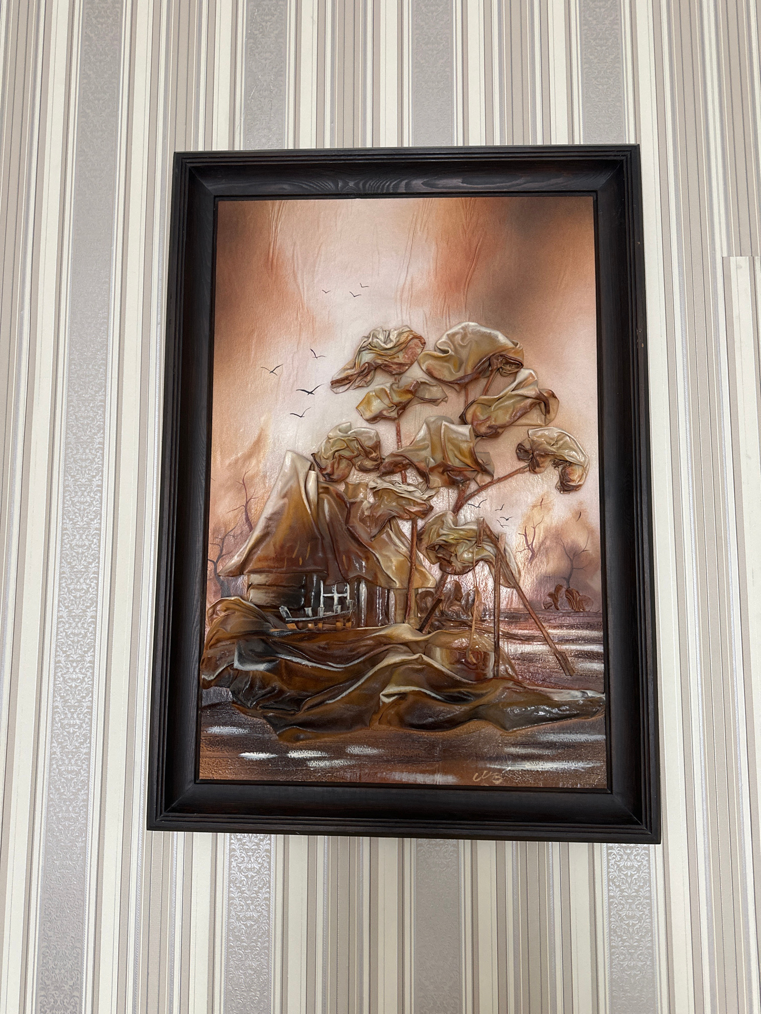 This image shows a framed relief artwork depicting a rustic scene with a thatched-roof house, stylized trees, and birds in flight, all rendered in warm brown and metallic tones. The artwork is hung on a wall covered with vertically striped wallpaper featuring alternating bands of cream, beige, and gray, with some stripes having a subtle embossed floral pattern. The lighting appears to be from above and slightly to the right, casting subtle shadows and highlighting the textured surface of the artwork. The scene is likely indoors, possibly in a residential setting, given the wallpaper. There is no indication of the specific location within the city of Minsk, Belarus.