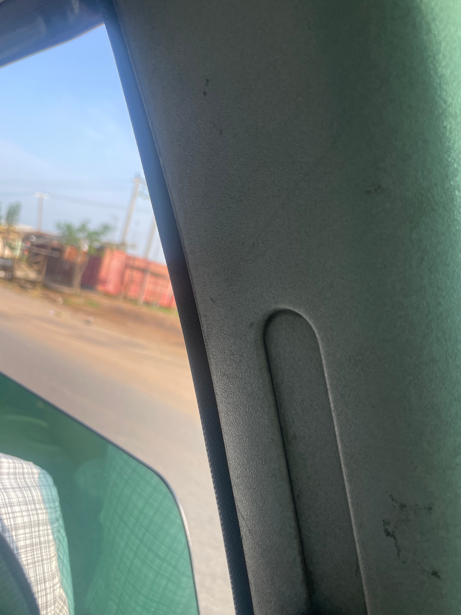 The image shows a view from inside a vehicle, looking out a window. The foreground is dominated by the interior of the car, specifically a grey textured surface that appears to be part of the car's pillar or dashboard. A green tint is visible on the lower portion of the window. Outside the window, a street scene in Wauro Jabbe, Nigeria, is depicted. The street is paved and appears to be a dusty road. On the left side of the road, there are several buildings, including what looks like shops or makeshift structures with corrugated metal roofs and some red shipping containers. A person wearing a checkered shirt is partially visible through the window on the left. The sky is clear and blue.