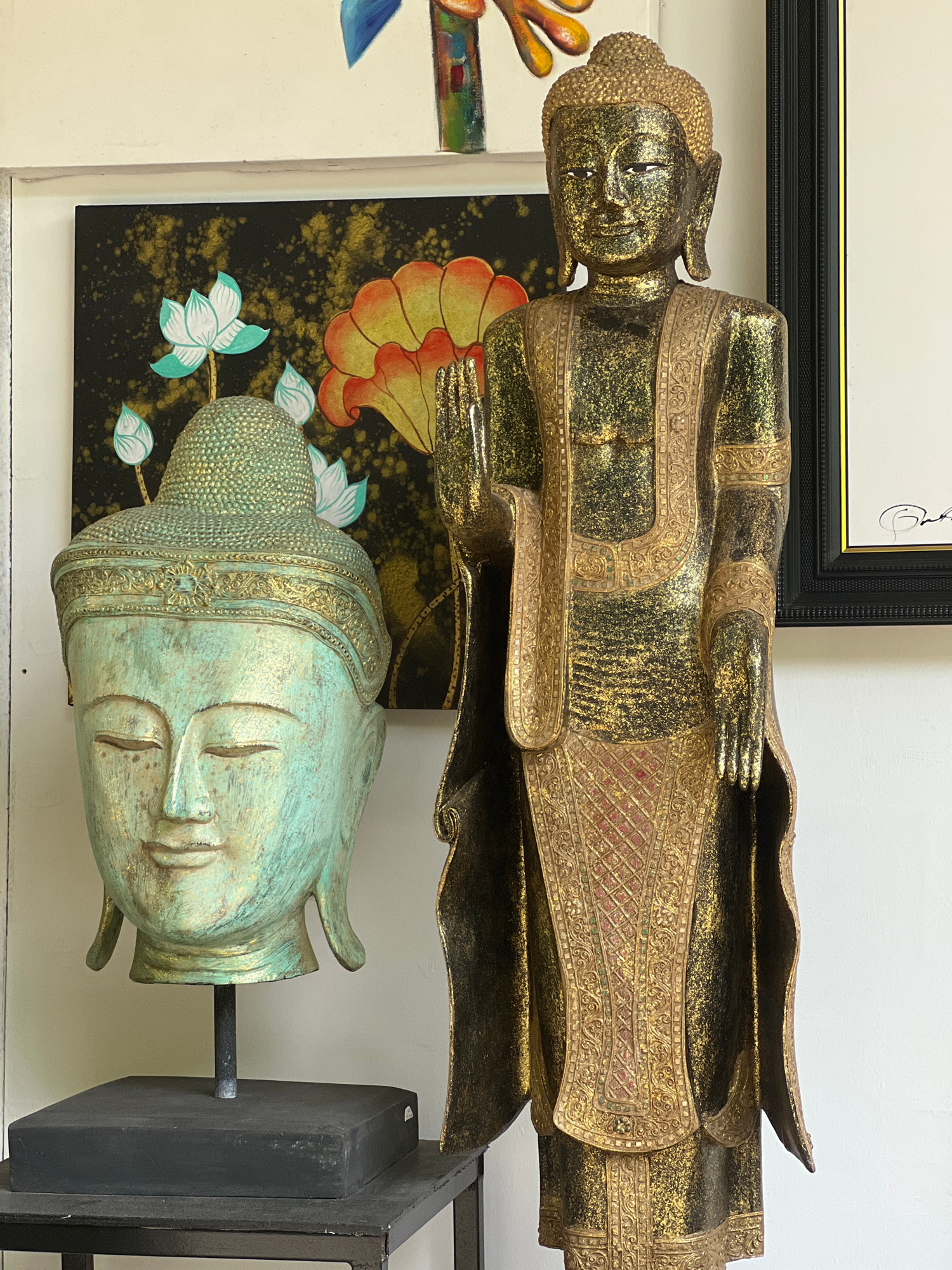 The image depicts a scene from Chalong, Thailand, featuring two Buddha statues and a piece of artwork. On the left is a large, teal-colored Buddha head mounted on a black square base. The head is adorned with gold accents and has closed eyes. Behind the head is a framed painting with a black background, featuring stylized lotus flowers in teal and orange hues with gold splatters. To the right of the Buddha head stands a tall, full-body Buddha statue, predominantly gold with black accents and intricate decorative patterns on its robes. The statue is positioned slightly in front of a framed artwork, possibly a document or print, with a black frame and white matting. The overall setting appears to be indoors, with a white wall as the backdrop.