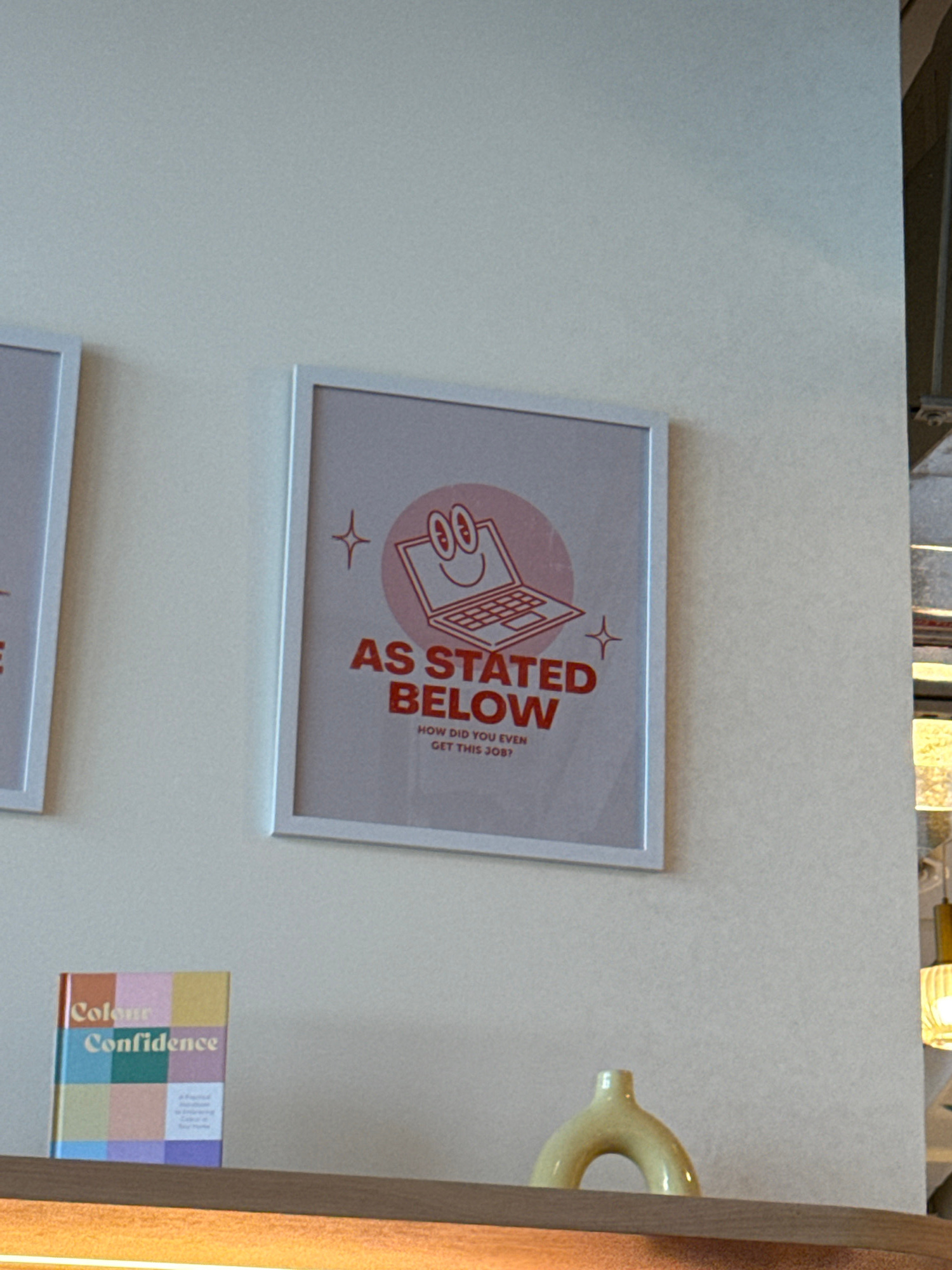 The image shows a framed artwork on a light-colored wall. The artwork features a graphic illustration of a laptop with a smiling face on the screen, surrounded by a pink circle. The text "AS STATED BELOW" is prominently displayed in red, with smaller text below that reads "HOW DID YOU EVEN GET THIS JOB?". Two small star-like graphics are also present on either side of the central illustration.
To the right of the framed artwork, a yellow, donut-shaped ceramic object is visible, mounted on the wall. Further to the right and slightly lower, a book with a colorful, checkered cover is placed on a shelf or surface. The visible text on the book reads "Colour Confidence" in large, bold letters. The overall scene appears to be an interior setting, possibly a home or an office, with warm lighting suggesting it could be daytime. The location context provided is Dubai, United Arab Emirates.