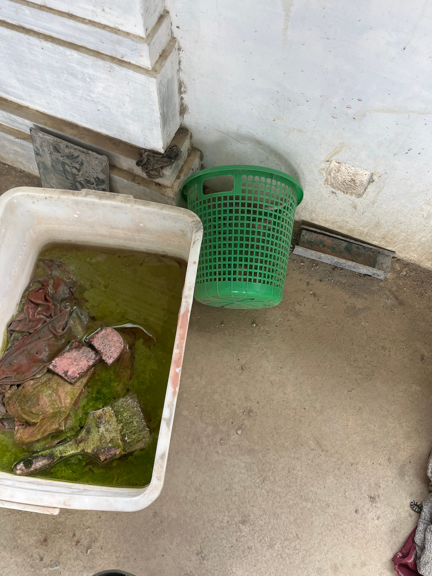 This media file captures an outdoor scene with a focus on objects, likely in a utilitarian or storage area. In the foreground, a large white rectangular tub is filled with murky green water. Submerged within the tub are several items, including what appears to be a paintbrush with a worn wooden handle, some pieces of reddish-brown material that might be fabric or brick fragments, and a clump of what looks like moss or algae.
To the left of the tub, a bright green plastic basket is partially visible, its grid-like structure evident. Behind the basket, a section of a rough, light-colored wall is visible, showing some wear and patches. To the right of the green basket, there's a narrow, metallic object, possibly a channel or trough, lying on the ground. The ground itself is a textured, sandy or dirt surface, with some small pebbles scattered around. In the upper right corner, a corner of what looks like patterned fabric or a blanket is draped over something, providing a hint of textiles in the scene.
The lighting suggests it is daytime, possibly overcast given the even illumination and lack of strong shadows. The overall impression is one of a somewhat unkempt or storage area, possibly a backyard, workshop, or an area where items are being cleaned or left to soak. The presence of the paintbrush and tub filled with water suggests an activity related to cleaning or art supplies, although the state of the water and the submerged items imply it might be a process that has been ongoing or is being neglected. No people are visible in the frame, and there is no discernible text. The location context is Benuaraja, Indonesia.