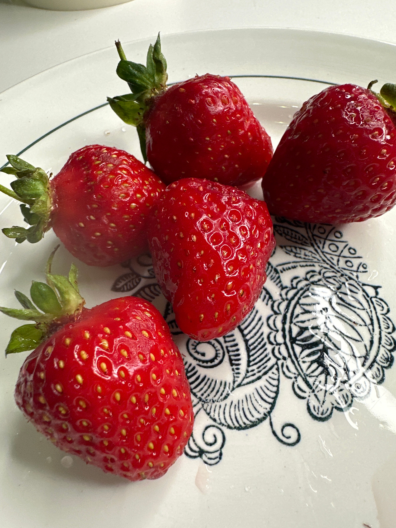 Five strawberries are arranged on a white plate with a black decorative pattern. The strawberries are bright red and appear fresh, with visible seeds and green stems. The plate has a dark green rim and is positioned on a light-colored surface.
