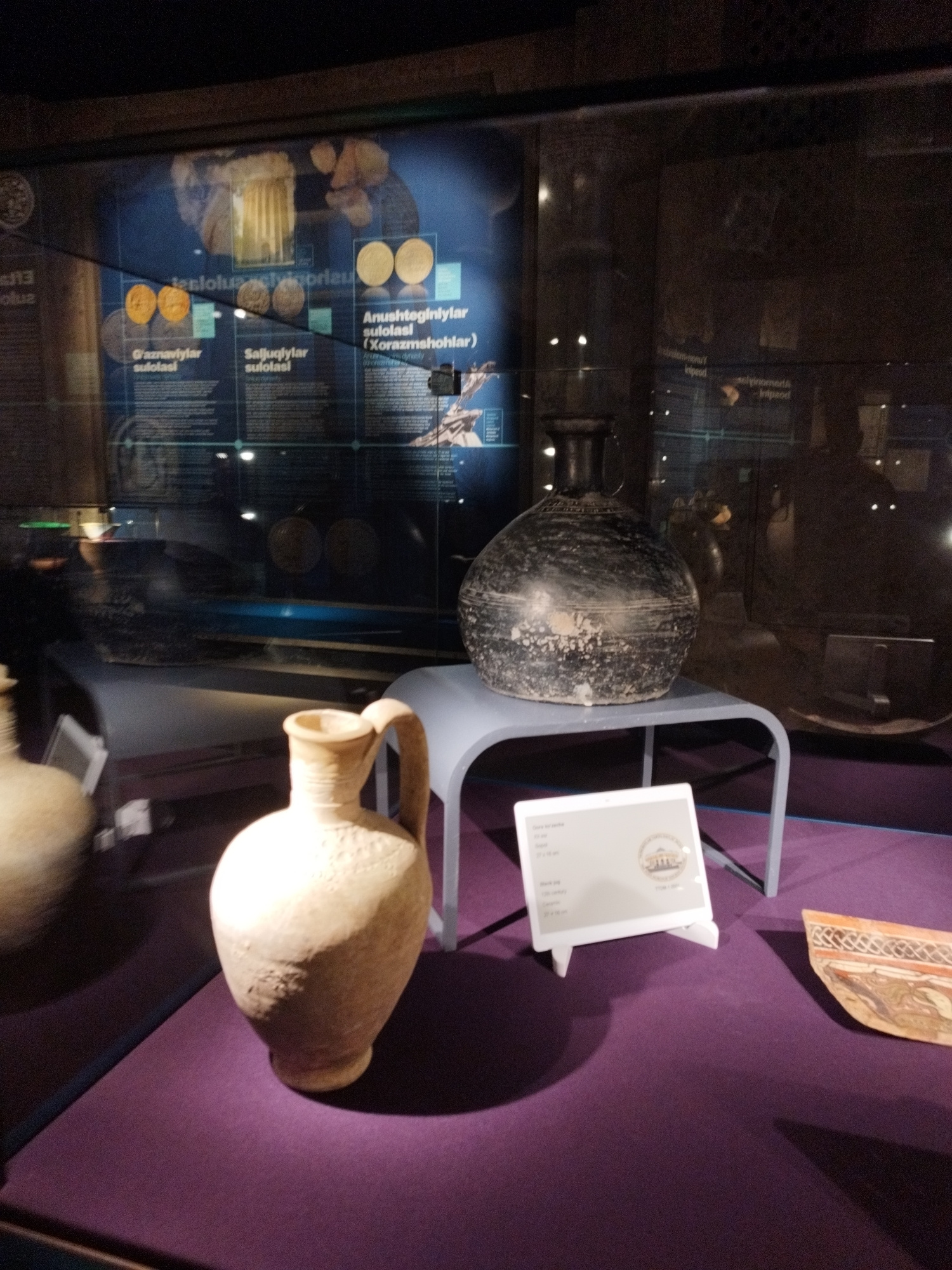 This media file shows a museum display featuring ancient artifacts. The primary subjects are two ceramic vessels: a light-colored jug with a handle and decorative patterns, and a dark, large, round vase with a narrow neck and two handles. Both are positioned on a purple surface within a glass display case.
In the background, a blue informational panel is visible, containing text in what appears to be a foreign language and images of coins. The text "G'aznaviylar sulolasi", "Saljuqiylar sulolasi", and "Anushteginlylar sulolasi (Xorazmshohlar)" is legible on this panel, suggesting a historical context related to dynasties and rulers. A digital display or tablet is also placed in front of the artifacts, showing information and a logo. A fragmented piece of pottery with intricate designs is visible to the right of the digital display.
The lighting suggests an indoor setting, likely a museum, with spotlights illuminating the artifacts. The overall scene conveys a sense of historical preservation and exhibition. The reflections on the glass case indicate the presence of external light sources and possibly viewers. No people are directly visible in the frame. The image appears to have been taken at eye-level.