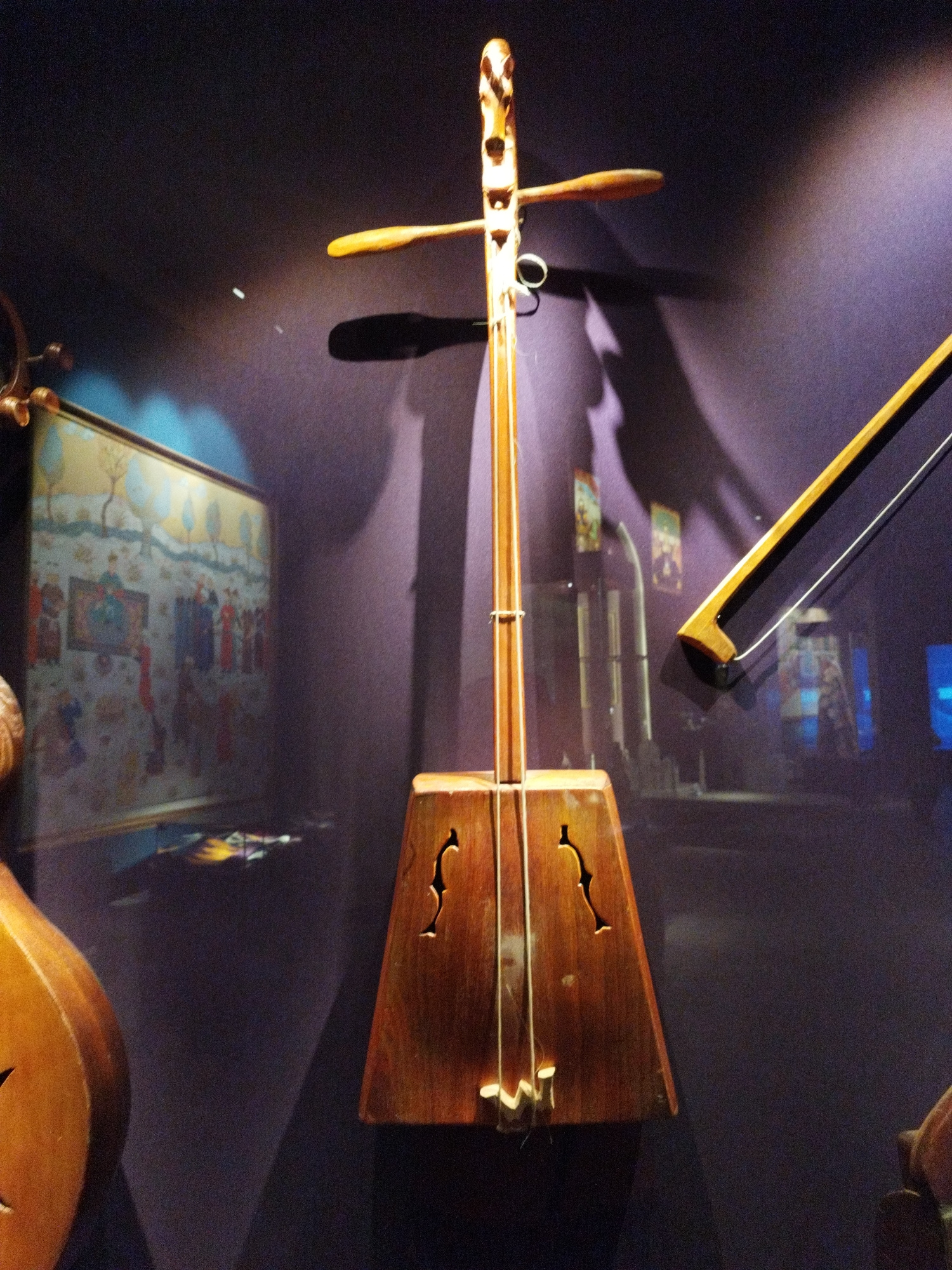 An upright, wooden stringed instrument with a long neck and a rectangular body is displayed against a dark purple background. The instrument has two strings that are visible, and the body features two stylized f-holes. A wooden bow with a visible string is positioned to the right of the instrument. To the left, partially visible, is another stringed instrument with a rounded body, also made of wood. In the background, a framed artwork depicts a scene with figures and trees. The lighting is dramatic, casting shadows and highlighting the instruments and artwork.