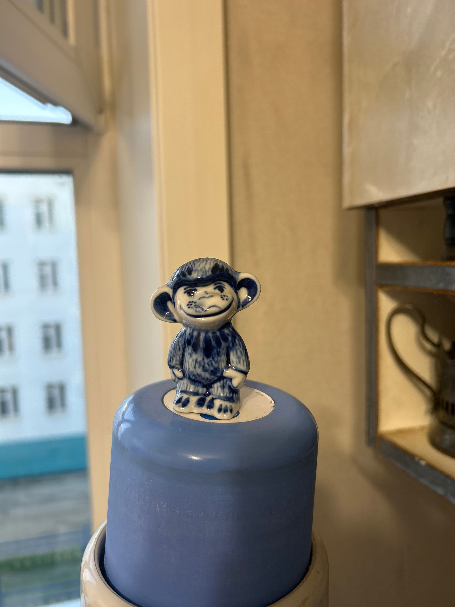 A close-up, eye-level, indoor photograph shows a small, blue and white ceramic figurine of a monkey standing on a matte, light blue cylindrical object. The monkey figurine is detailed with dark blue patterns that mimic fur and has a smiling expression. Its ears are large and prominent. The blue cylinder appears to be the lid of a larger object, with a cream-colored rim visible at the bottom of the frame.
In the background, out of focus, a window reveals a building with multiple windows, suggesting an urban environment. The light appears to be natural, coming from the window. The overall lighting is soft. The background wall to the right is a neutral, warm tone, and a shelf with a dark, metallic object (possibly a kettle or pitcher) is also visible but blurred.
The photograph was taken in Yakutsk, Russia, although the specific location within the city is not discernible from the image itself.
