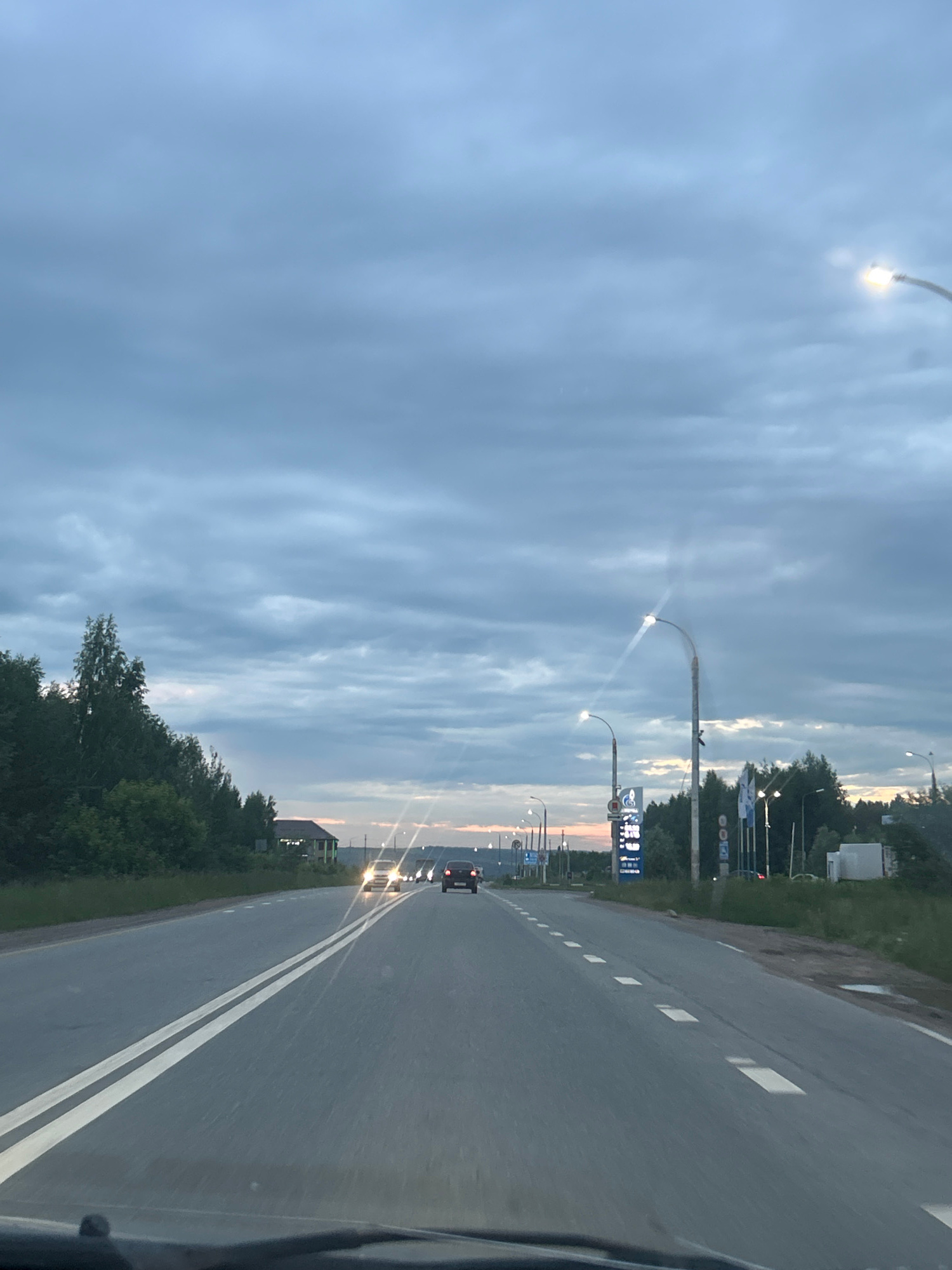 The image shows a perspective from inside a vehicle, looking forward along a two-lane asphalt road. The sky is cloudy and appears to be twilight. On the left side of the road, there are trees and bushes. On the right side of the road, there are streetlights, a gas station with a price sign, and some buildings in the distance. Several cars are visible on the road, some with headlights on. The road has double solid white lines marking the center. The scene is likely on the outskirts of a populated area, given the presence of a gas station and the open landscape beyond the road.