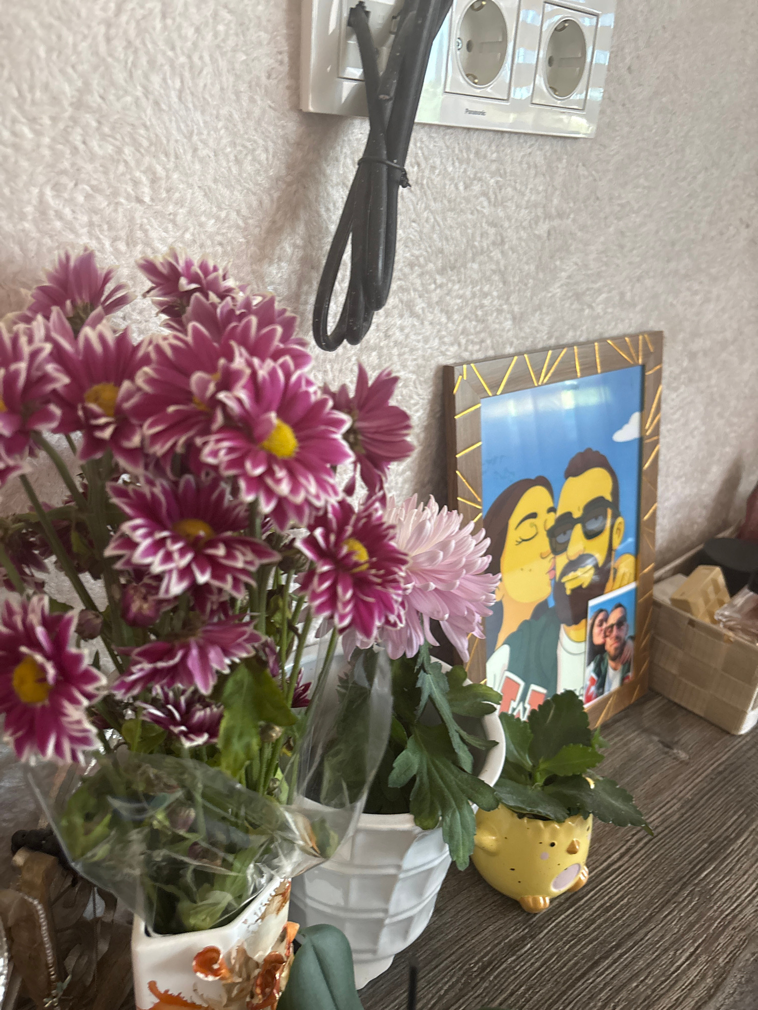 This is a medium-shot, eye-level, indoor photograph taken in Bulgarovo, Bulgaria. The scene depicts a shelf with several decorative items. In the foreground, a bouquet of purple and white chrysanthemums is partially wrapped in clear plastic, arranged in a white vase with an orange, nature-themed pattern. To the right of the flowers, a framed digital illustration of a couple kissing is displayed. The couple is depicted with yellow skin tones and is set against a blue sky with a white cloud. Below the illustration, a small framed photograph shows a real couple. Further to the right, a small yellow ceramic chick-shaped planter holds a green plant. In the background, a white electrical outlet with two sockets is visible on a textured, off-white wall, with a black electrical cord hanging from it. The shelf itself is made of wood with a visible grain pattern.