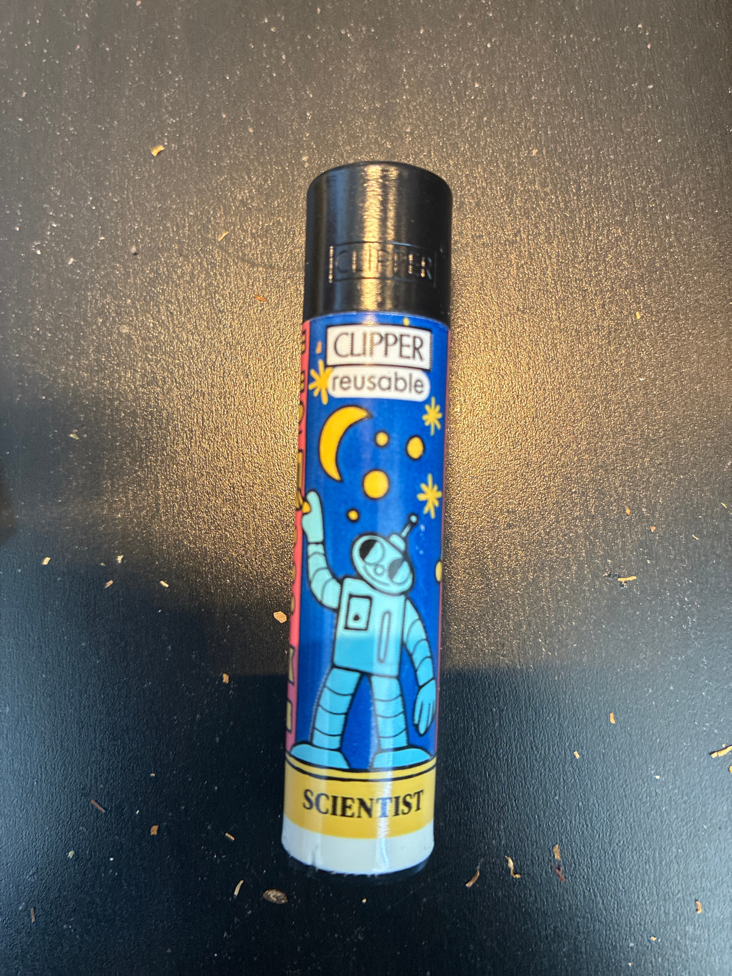 The image shows a Clipper lighter with an astronaut design. The lighter is black with a blue sleeve featuring a cartoon astronaut, a moon, and stars. The word "CLIPPER" is visible at the top of the sleeve, followed by "reusable" and "SCIENTIST" at the bottom. The lighter is placed on a dark, textured surface, possibly a table, with some small debris scattered around it. The background is out of focus, suggesting the image was taken from a close-up perspective. The image does not provide any information about its location in the city of l'Hospitalet de Llobregat, Spain.