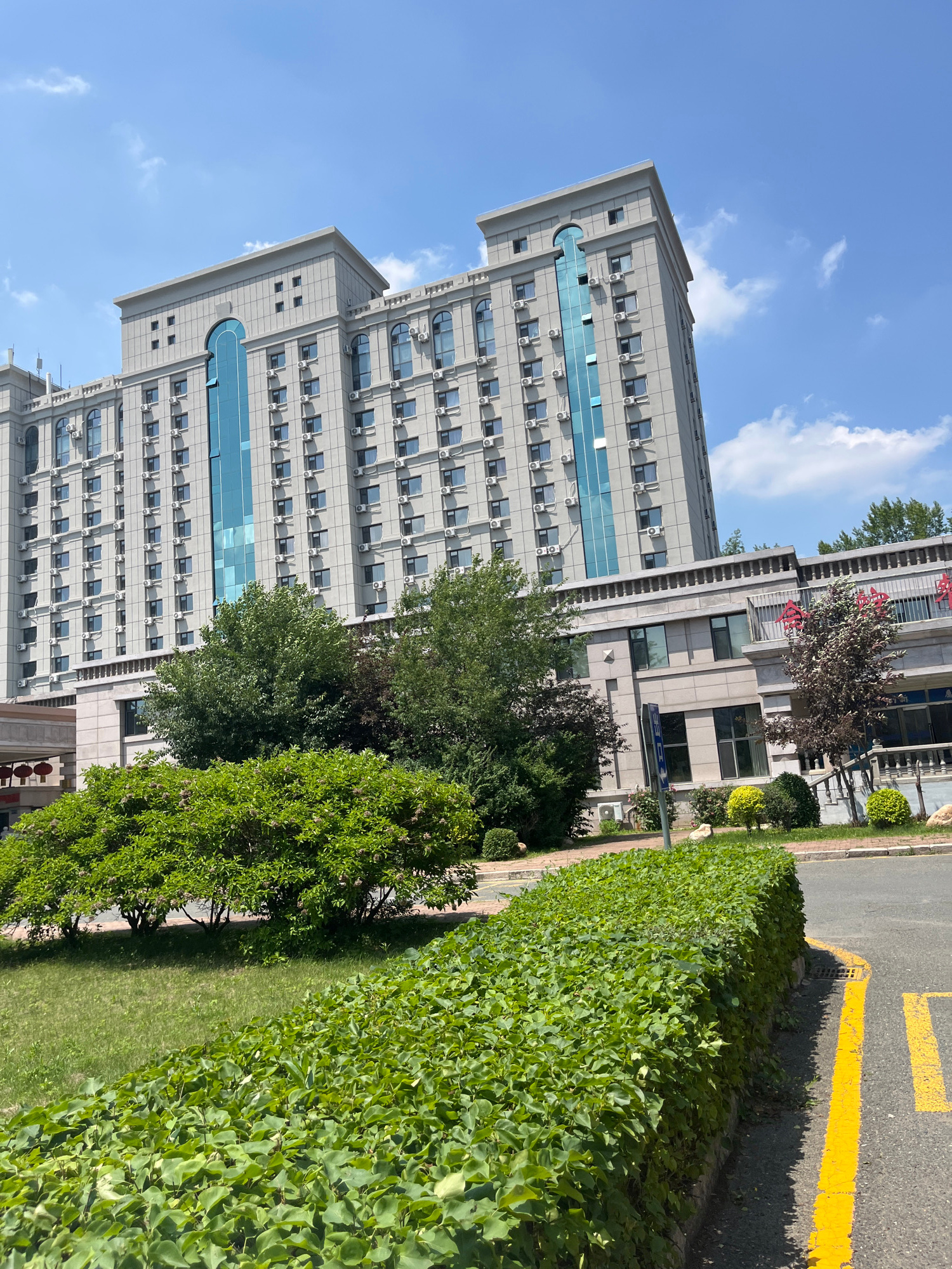 The image shows a modern building with a light-colored facade, featuring rows of windows and air conditioning units. The building has a prominent central section with blue glass accents. In front of the building, there are lush green trees and manicured bushes, creating a landscaped area. A paved road with yellow markings is visible in the foreground, bordering the greenery. The sky above is blue with scattered white clouds, indicating a clear day. The scene appears to be a typical urban environment with commercial or residential buildings and green spaces. Based on the architectural style and general appearance, this location could be a hotel, office building, or an educational institution within Changchun City, China.