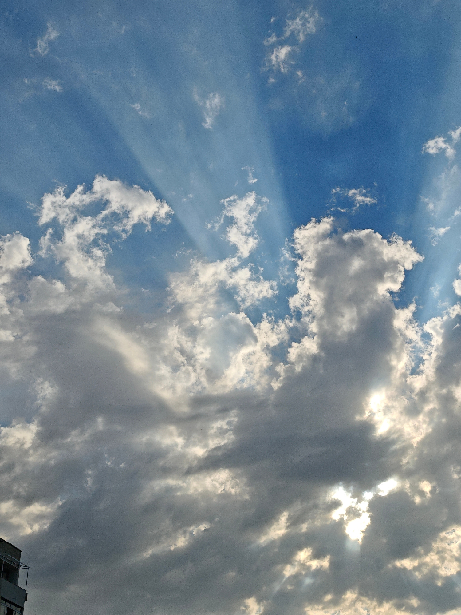 The image depicts a dramatic sky with crepuscular rays piercing through clouds. The sky is a vibrant blue, with fluffy white clouds scattered throughout, interspersed with darker, more ominous-looking clouds. The sun is partially obscured by these clouds, creating a halo effect and highlighting the beams of light. The lower left corner of the image shows the top portion of a building, suggesting an urban setting. The scene appears to be a natural phenomenon observed from a city. There are no people or discernible actions taking place. The image is a vertical shot looking upwards at the sky.