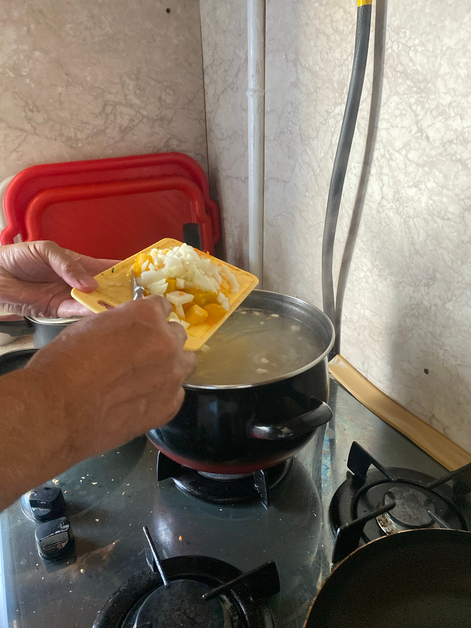 The image depicts a close-up, eye-level view of a person's hands adding chopped vegetables to a pot of simmering liquid on a gas stove. The vegetables, appearing to be onion and possibly bell pepper or squash, are on a small yellow cutting board, from which they are being scraped into the pot using a utensil. The pot is black with red accents, resting on a black gas stovetop with two burners visible. To the left, stacked red plastic trays are partially visible. The background features a tiled wall with a beige, marble-like pattern. A dark, flexible tube, possibly a gas line, extends vertically along the right side of the frame. The lighting is moderate, casting some shadows. The scene appears to be in a domestic kitchen setting.