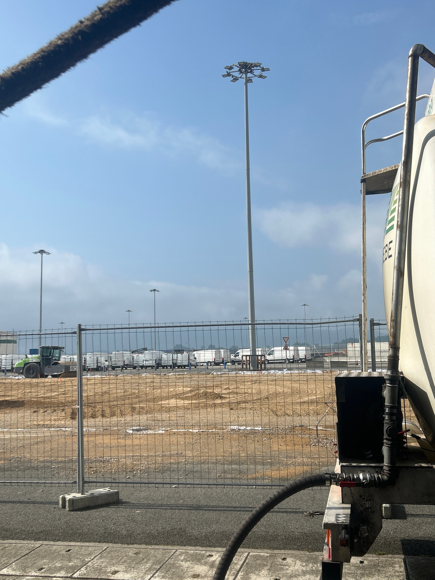 The image depicts a construction site in Santander, Spain, under a clear blue sky with scattered clouds. In the foreground, a portion of a large white tank truck is visible on the right, with a black hose attached to its side. A metal railing and a ladder are also part of the truck's structure. A chain-link fence runs across the midground, separating the foreground from the construction area. Behind the fence, a green construction vehicle, possibly a roller, is parked on a patch of disturbed earth. Further back, a row of white vans or small trucks is lined up. Tall light poles are prominent features in the background, illuminating the expansive site. The ground in the foreground appears to be asphalt, with concrete slabs and edging visible. The overall scene suggests ongoing construction or industrial activity at a port or large facility.