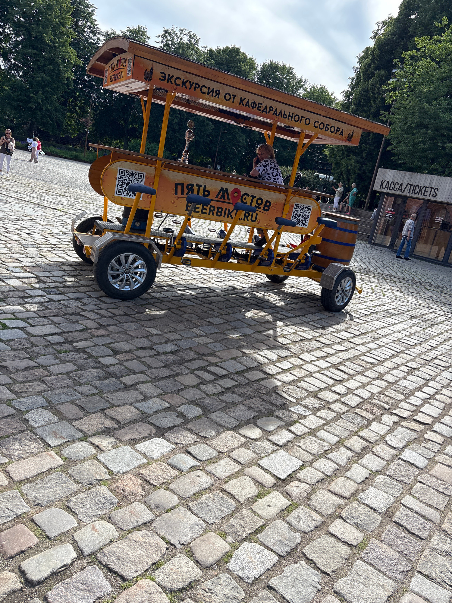 This image shows a yellow pedal-powered vehicle, designed to carry multiple passengers, parked on a cobblestone plaza. The vehicle is branded with Russian text, including "ЭКСКУРСИЯ ОТ КАФЕДРАЛЬНОГО СОБОРА" which translates to "Excursion from the Cathedral". Another prominent text on the vehicle reads "ПЯТЬ МОСТОВ BEERBIKE.RU". In the background, a building with a sign reading "КАССА/TICKETS" is visible. Several people are walking in the plaza and near the building. The setting appears to be a public square, likely a tourist area, given the presence of the excursion vehicle and ticket booth. The architecture and cobblestone paving suggest a historical European city. This specific vehicle and the Cathedral mentioned are associated with the city of Kaliningrad, Russia, specifically in the vicinity of the reconstructed Königsberg Cathedral.